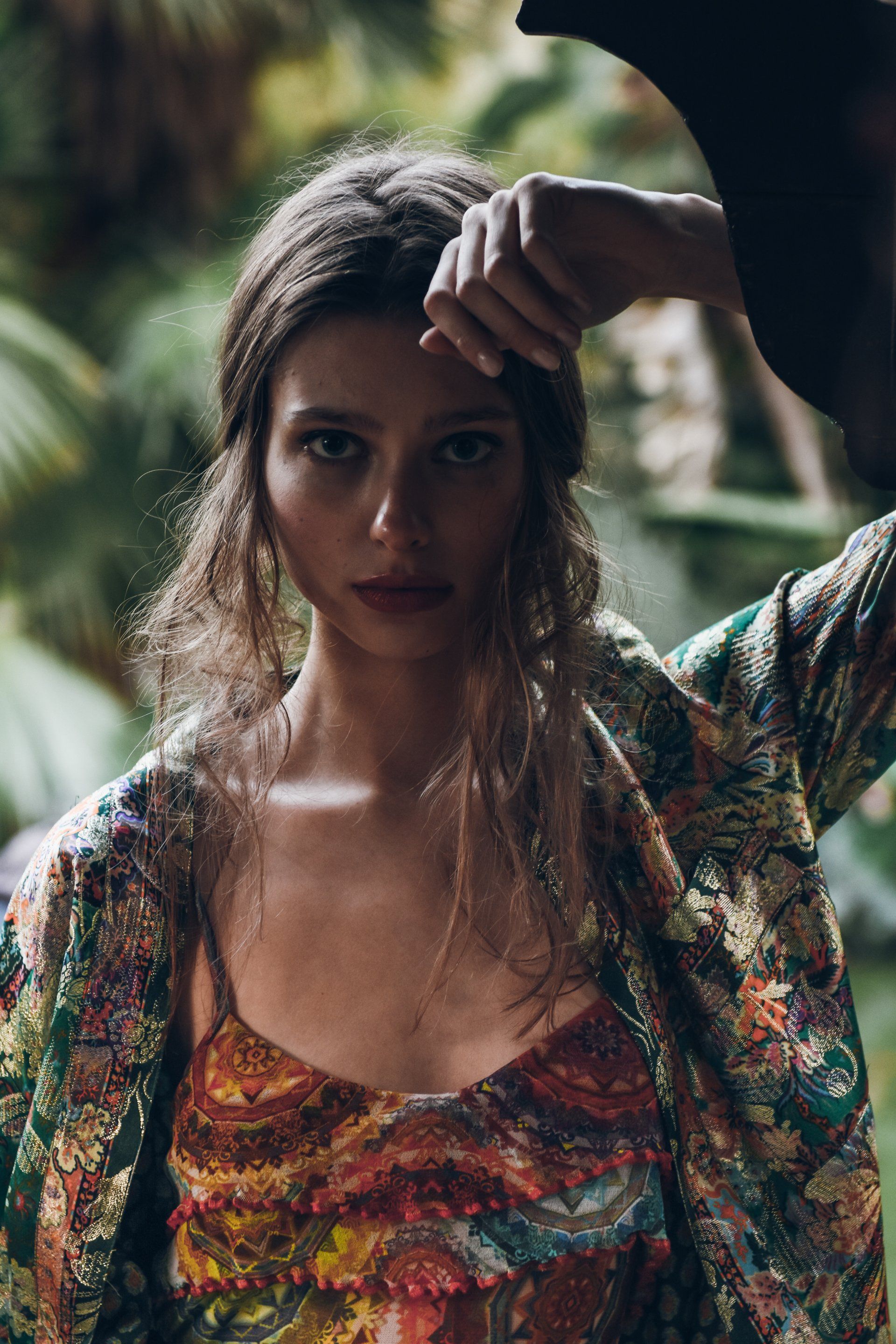 Frau in bunt bedrucktem Oberteil und Jacke, Hand auf dem Kopf, im Freien mit Laub.