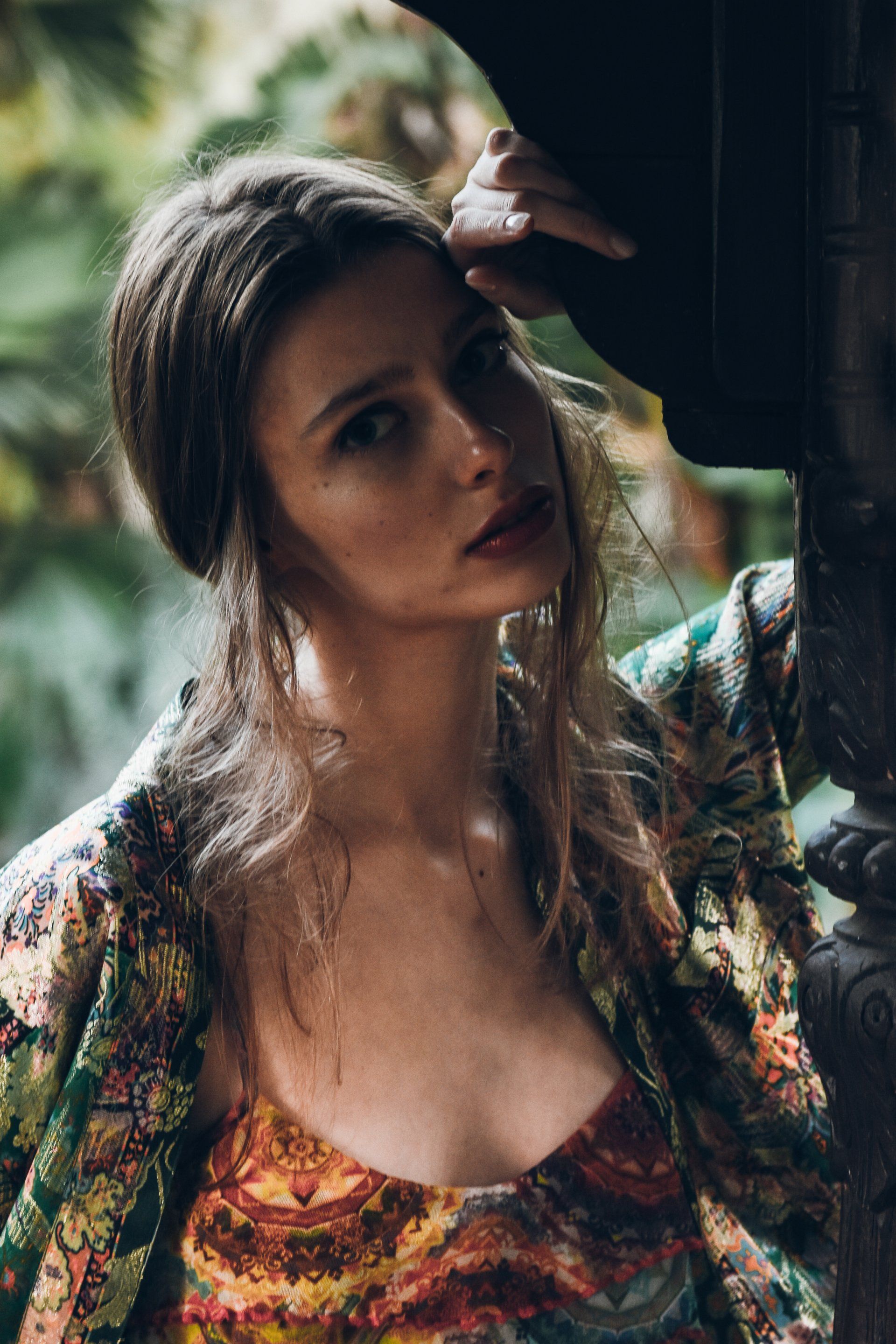 Frau mit welligem Haar, gemusterter Jacke, lehnt an verzierter Struktur und blickt den Betrachter an.