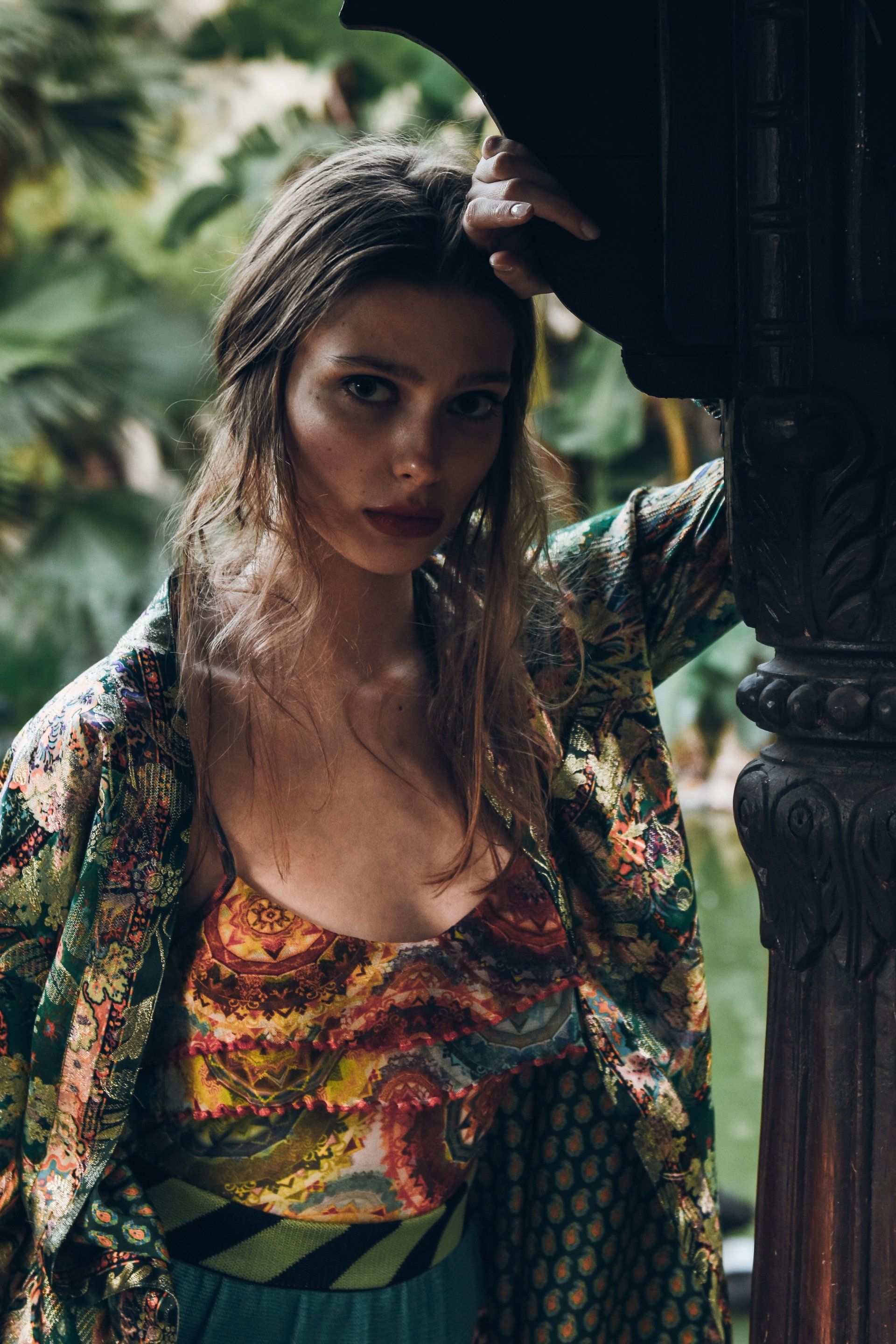 Frau in bunt gemusterter Kleidung lehnt im Freien an einer geschnitzten Säule.