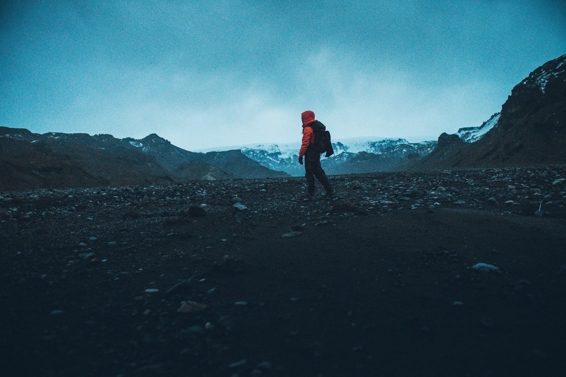 Wanderer in roter Jacke steht auf dunklem, felsigem Boden, im Hintergrund Berge unter einem bewölkten Himmel.
