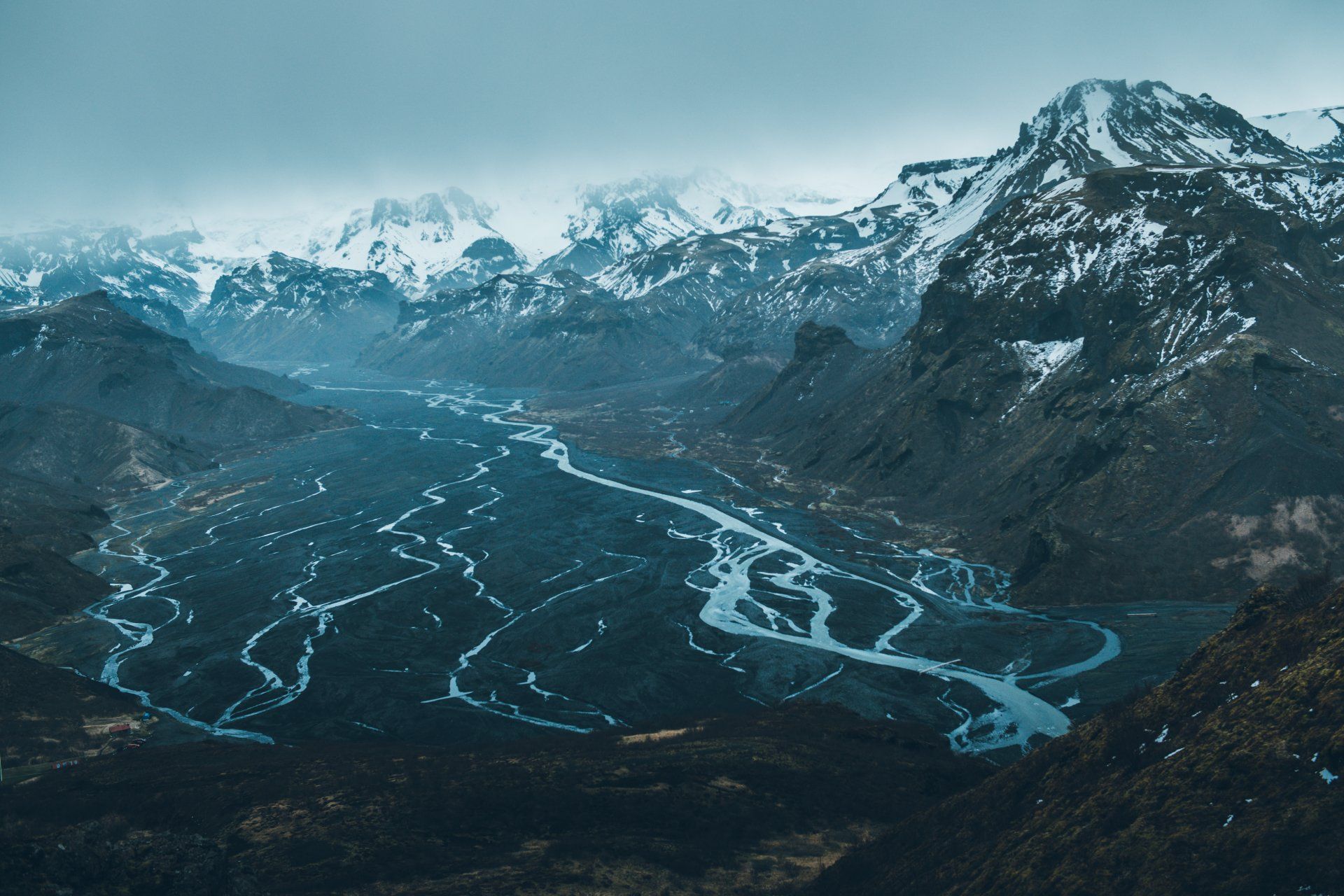 Berge mit schneebedeckten Gipfeln überblicken ein Flusstal mit einem dunklen, mäandernden Fluss und bewölktem Himmel.