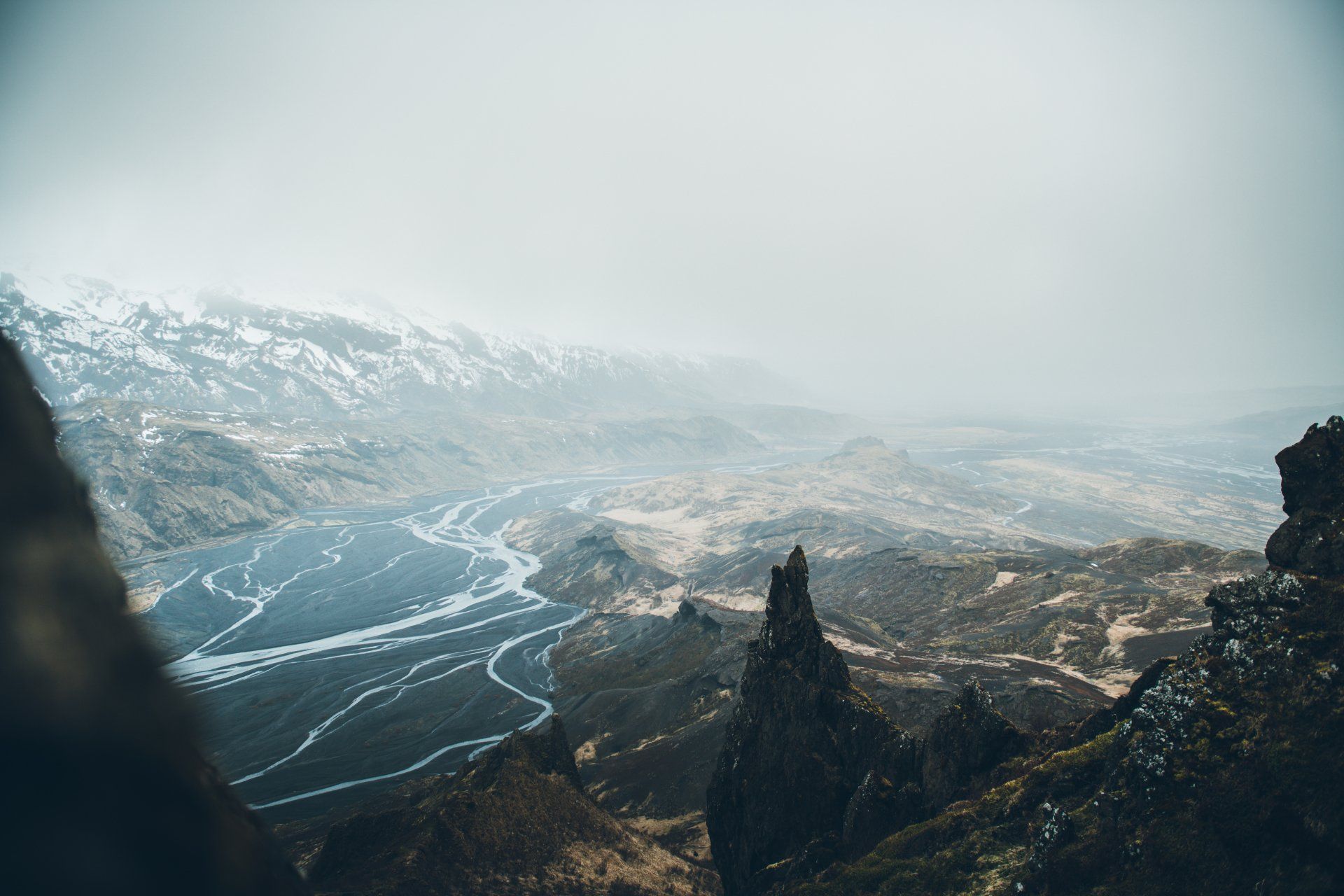 Blick auf einen Fluss, der sich durch ein Tal unter einer wolkigen, schneebedeckten Bergkette schlängelt.