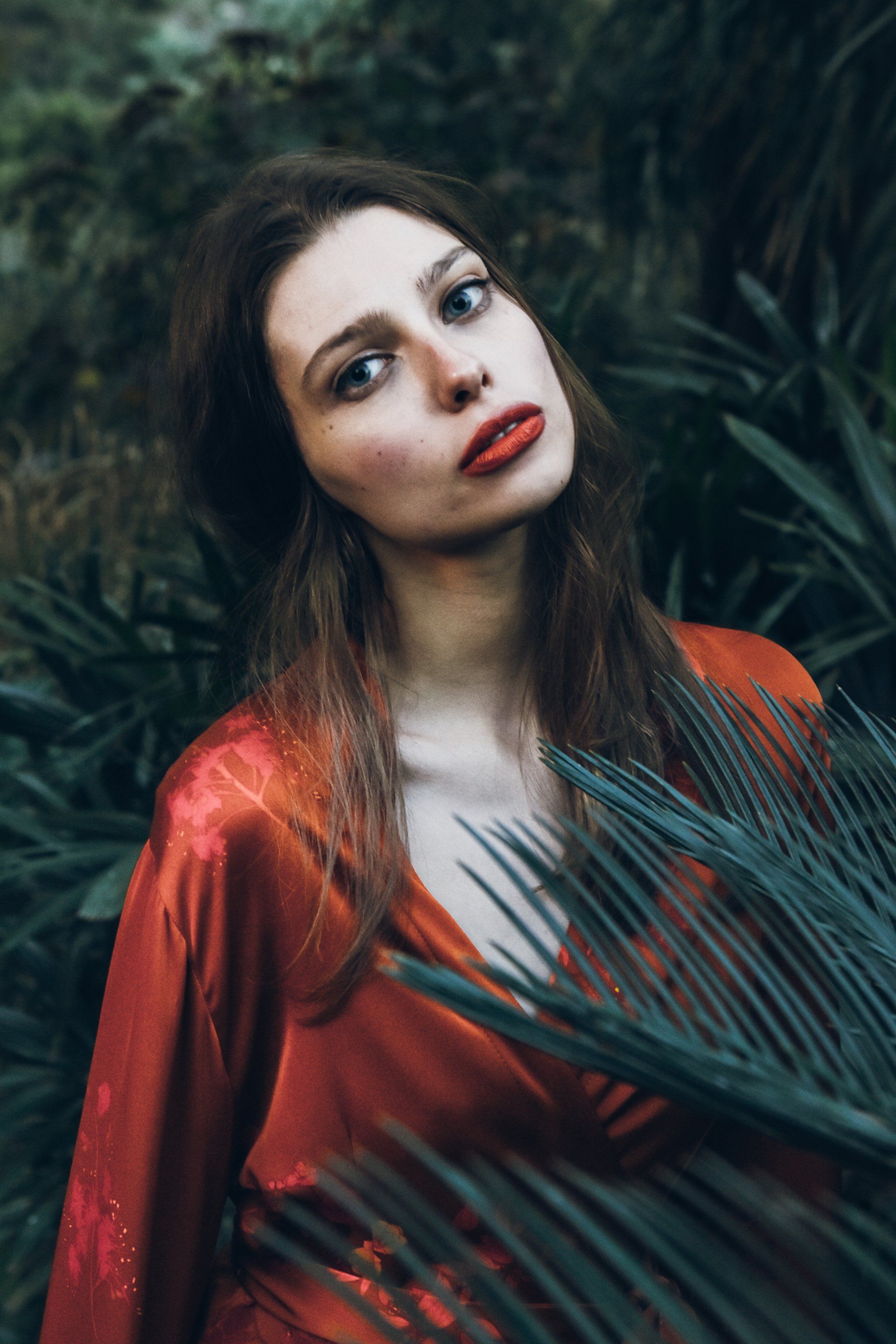 Frau im roten Kleid mit rotem Lippenstift, umgeben von grünen Blättern; im Freien.