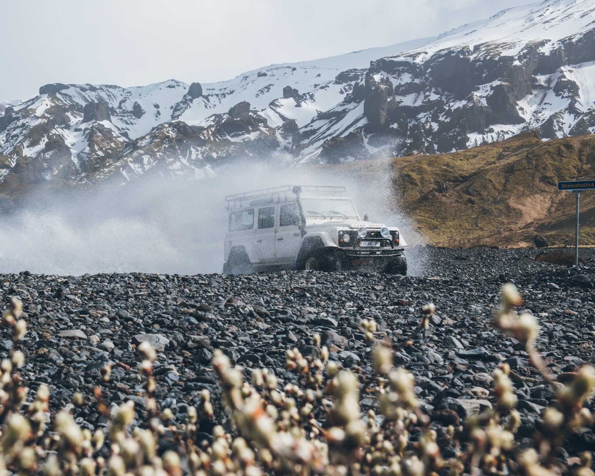 Weißer SUV durchquert einen Bach, Wasser spritzt. Im Hintergrund schneebedeckte Berge, im Vordergrund felsig.
