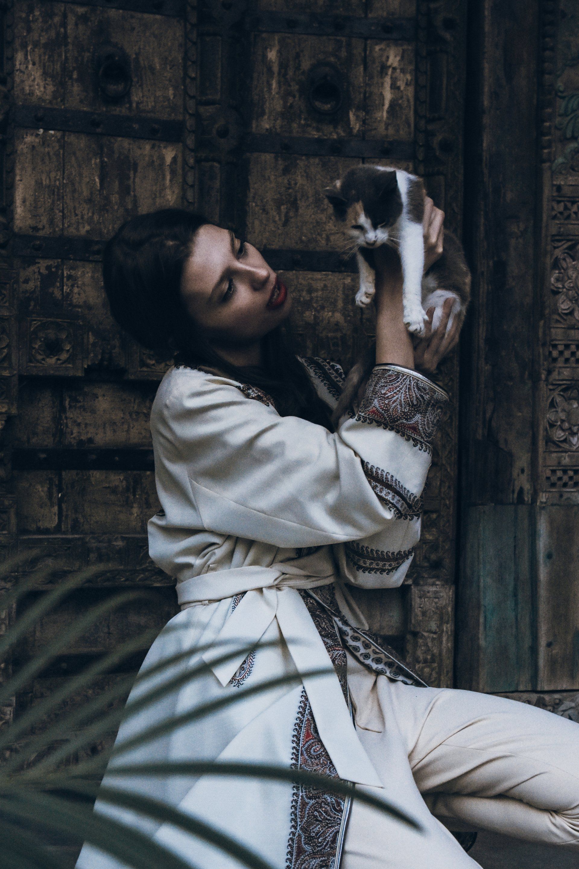 Frau im weißen bestickten Outfit hält ein Kätzchen und schaut es an. Alter Holztürhintergrund.