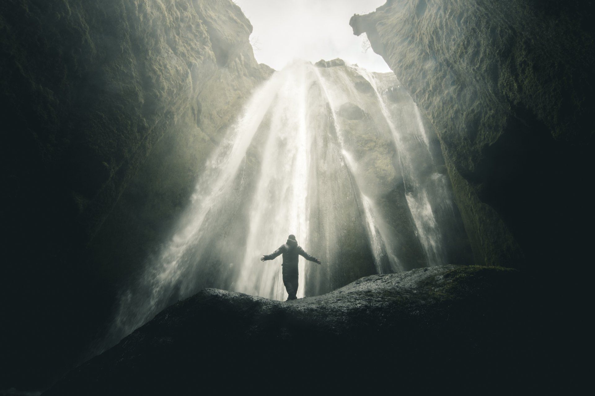 Person mit ausgestreckten Armen steht vor einem Wasserfall in einer dunklen Höhle.