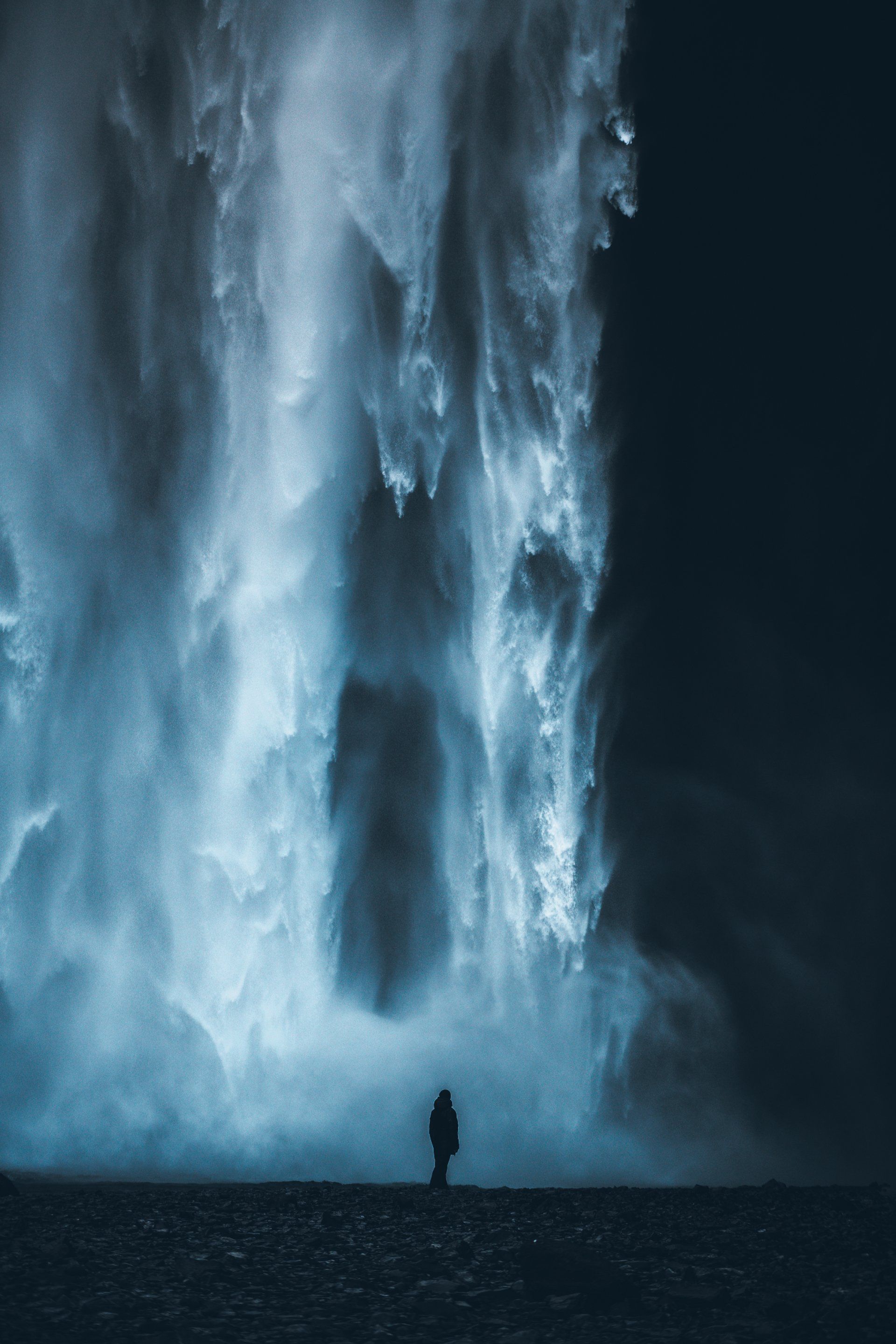 Silhouette einer Person, die nachts vor einem hoch aufragenden blauen Wasserfall steht.