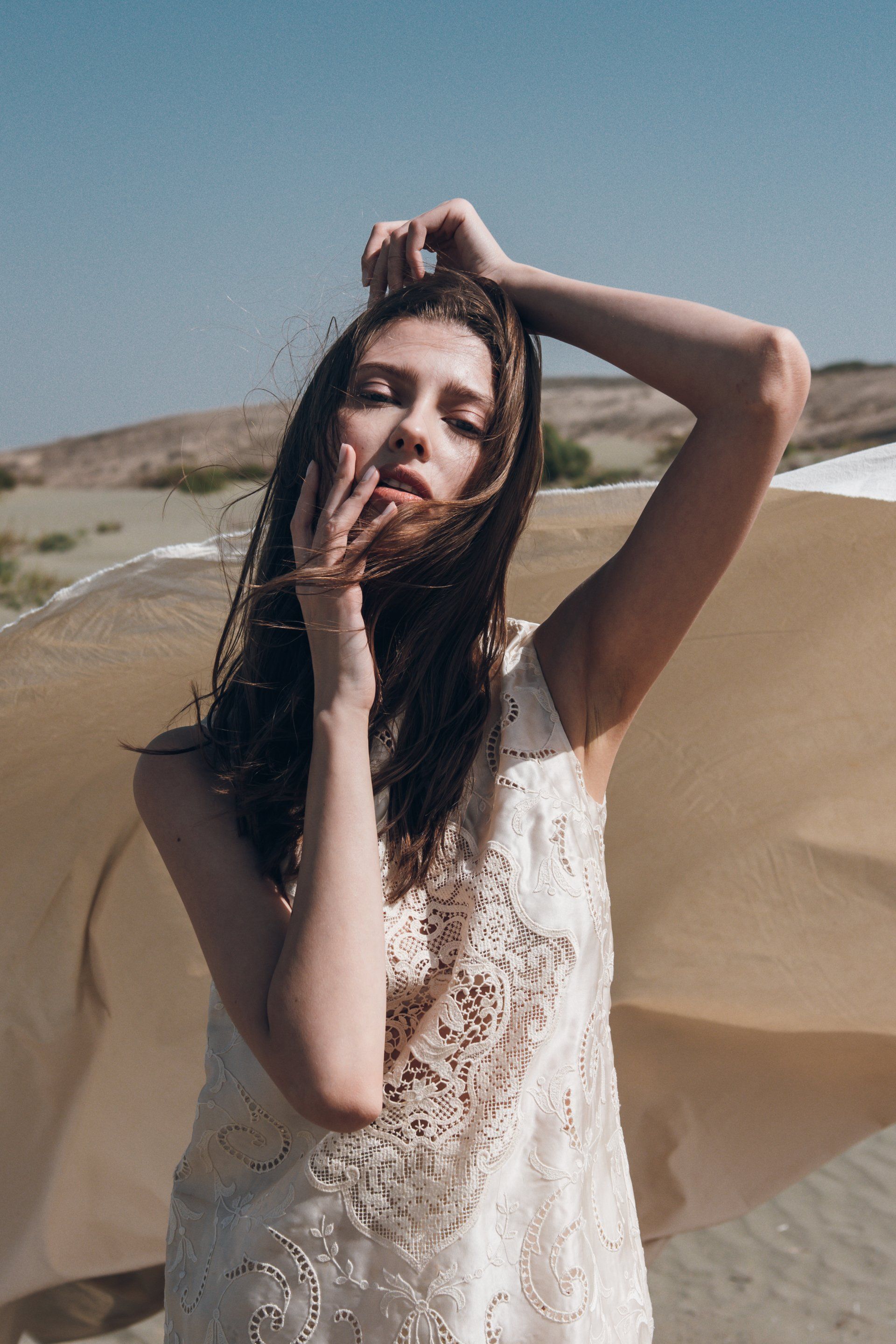 Frau im weißen Kleid, Arme erhoben, posiert in der Wüste, hellbrauner Stoff weht im Wind, blauer Himmel.