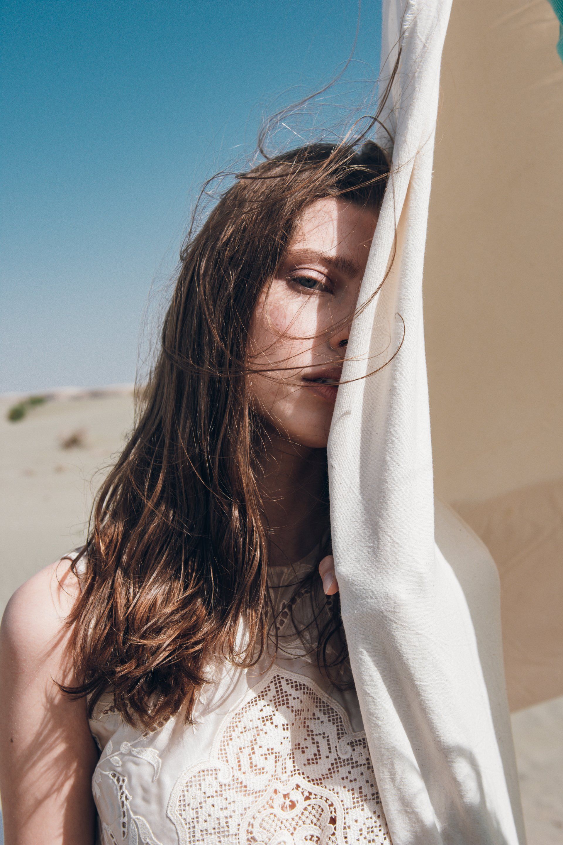 Frau mit langen braunen Haaren, deren Gesicht teilweise mit weißem Stoff bedeckt ist, steht draußen im Sonnenlicht.
