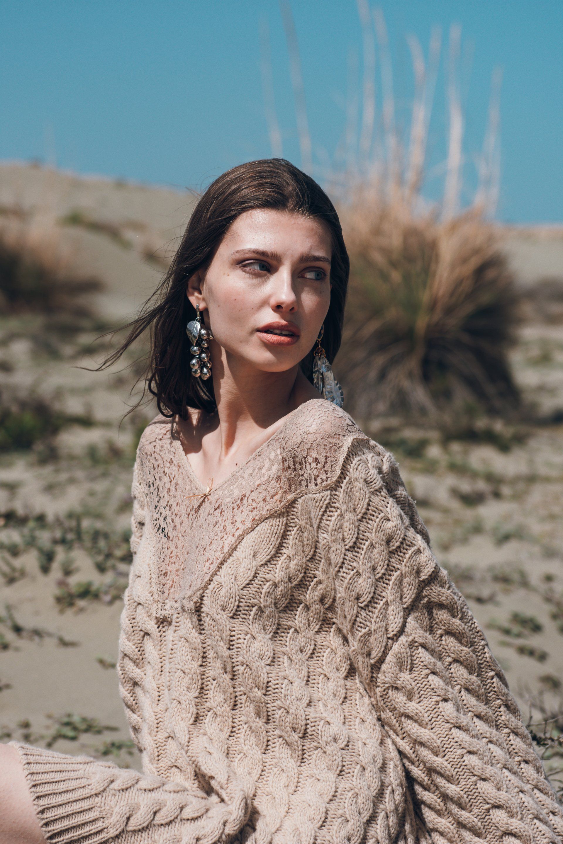Frau in beigem Zopfmusterpullover auf einer Sanddüne, Blick zur Seite.