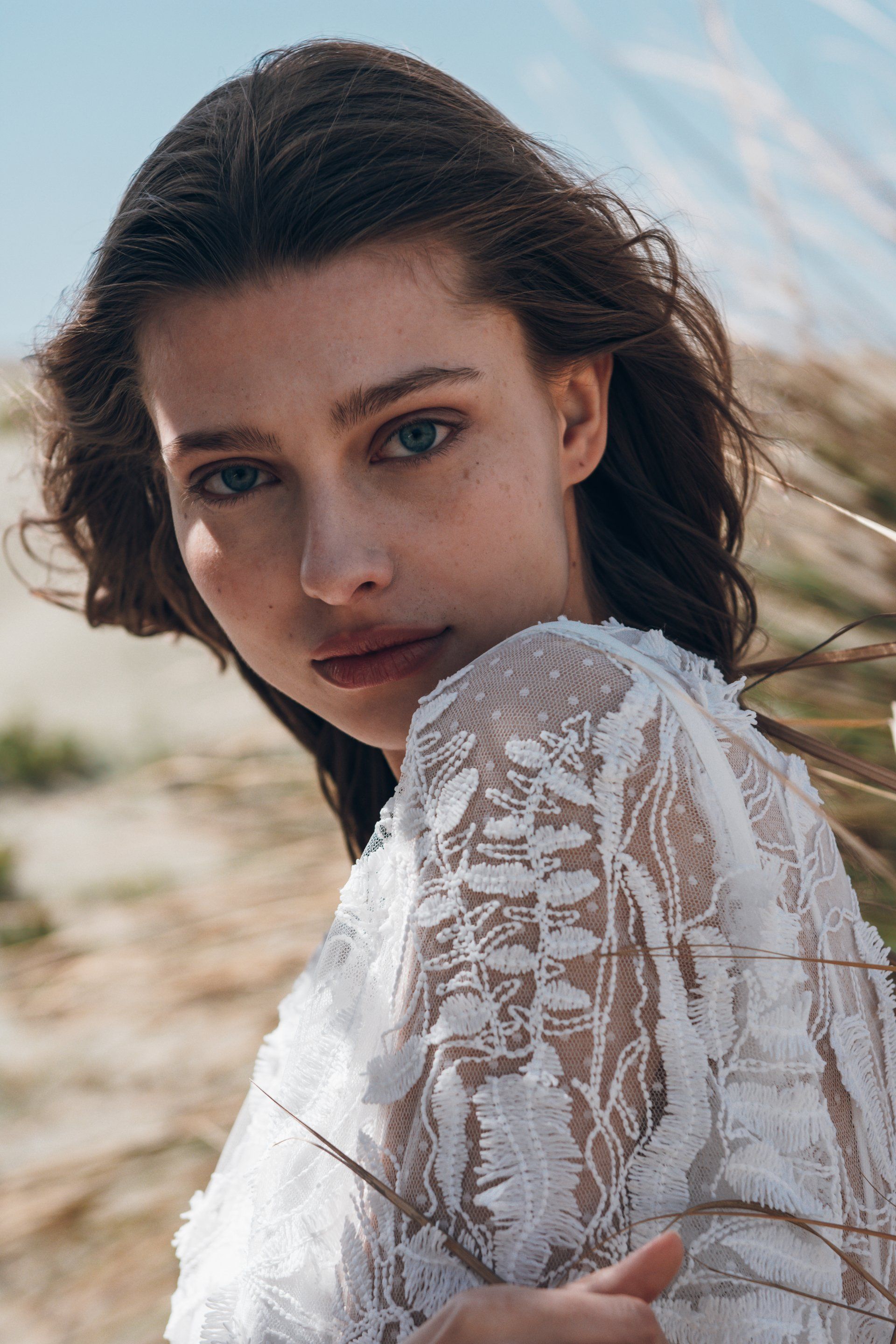 Frau im weißen Spitzenkleid posiert an einem Strand und blickt mit sanftem Gesichtsausdruck in die Kamera.