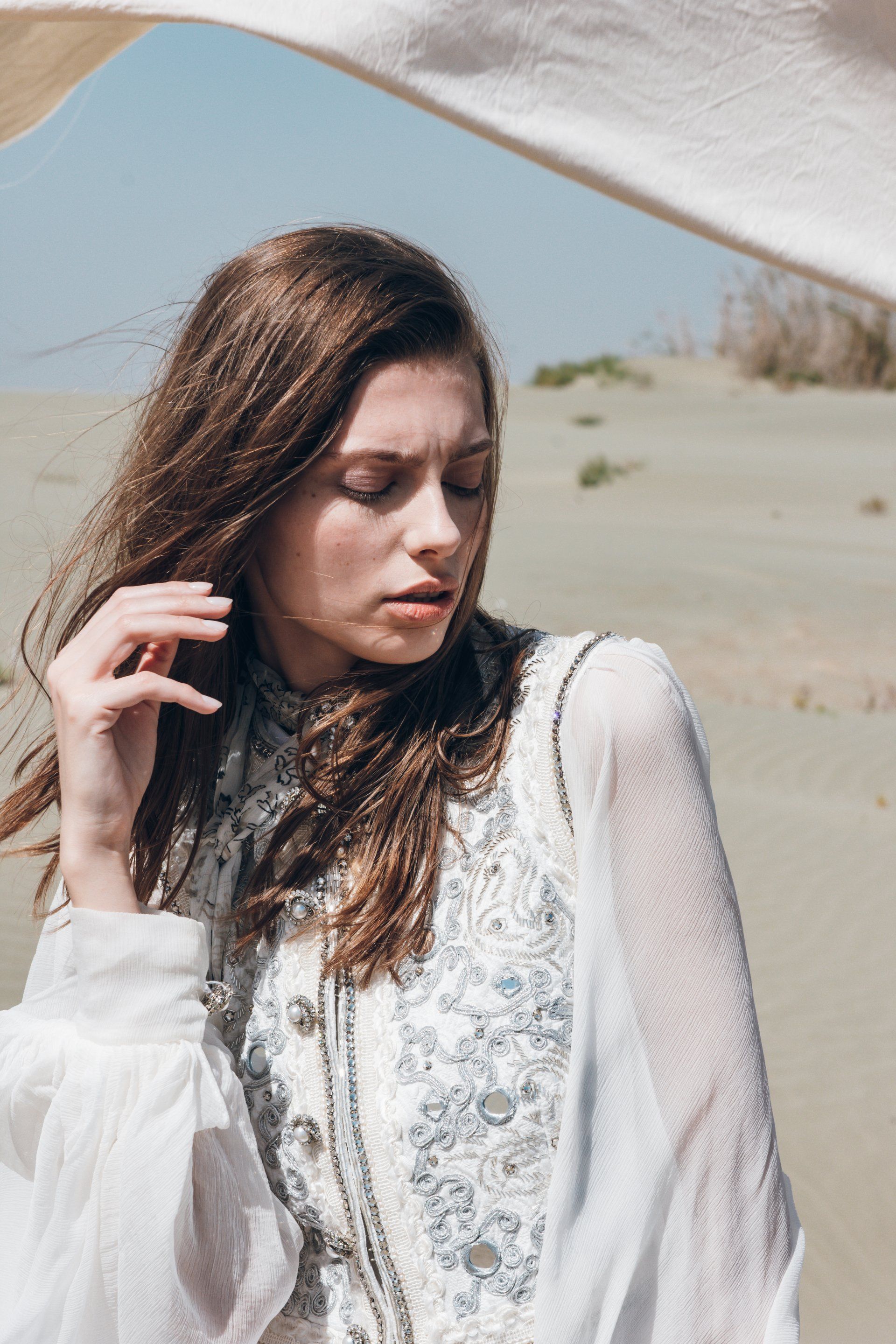 Frau in weißem Kleid mit kunstvollen Details, Augen geschlossen, Hand im Haar, posiert auf Sand.