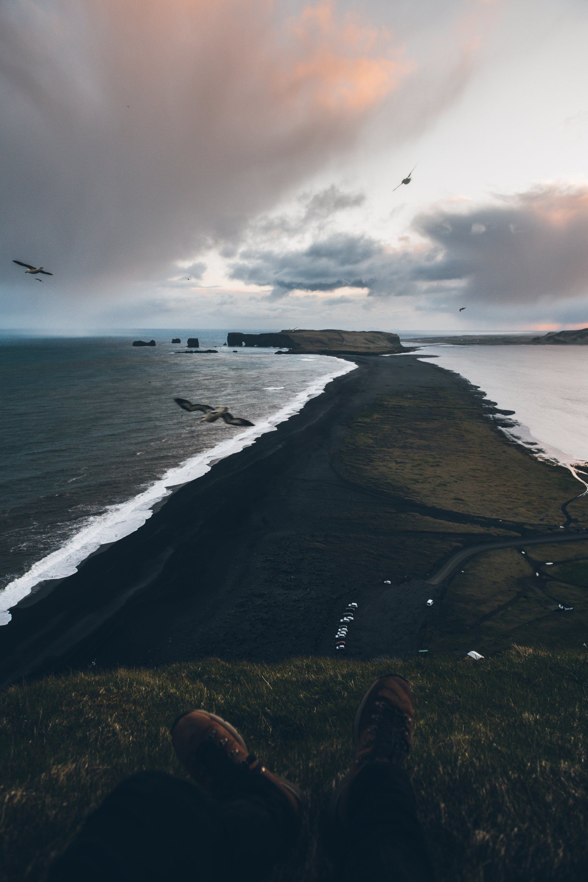 Schwarzer Sandstrand, der den Ozean unter bewölktem Himmel teilt. Im Vordergrund die Füße einer Person.