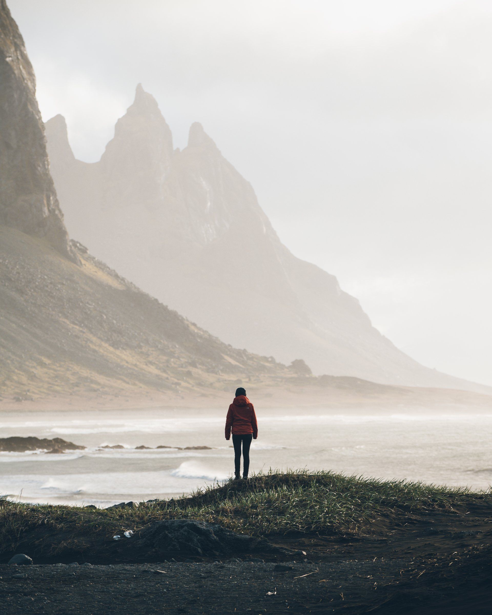 Eine Person in einer roten Jacke steht an einem Ufer und blickt auf hoch aufragende Berge in einer nebligen Umgebung.