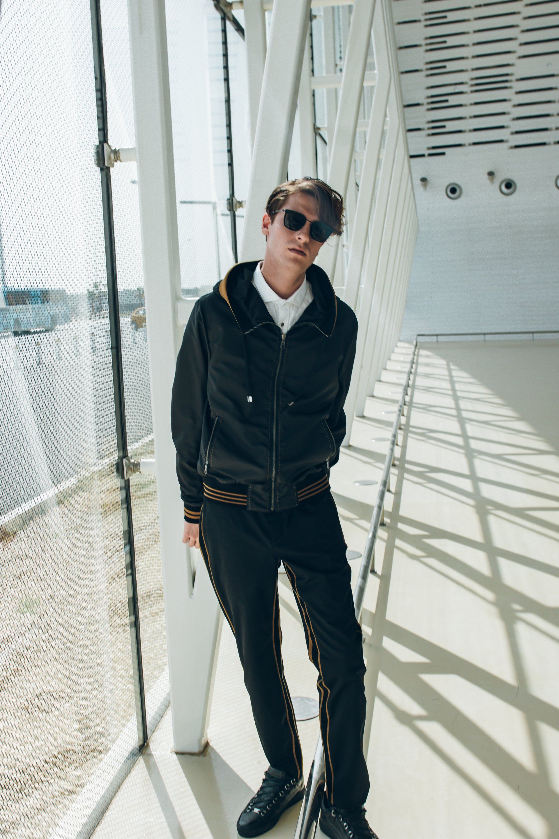 Ein Mann mit Sonnenbrille, schwarzer Jacke und Hose steht an einem Fenster in einem modernen Gebäude.