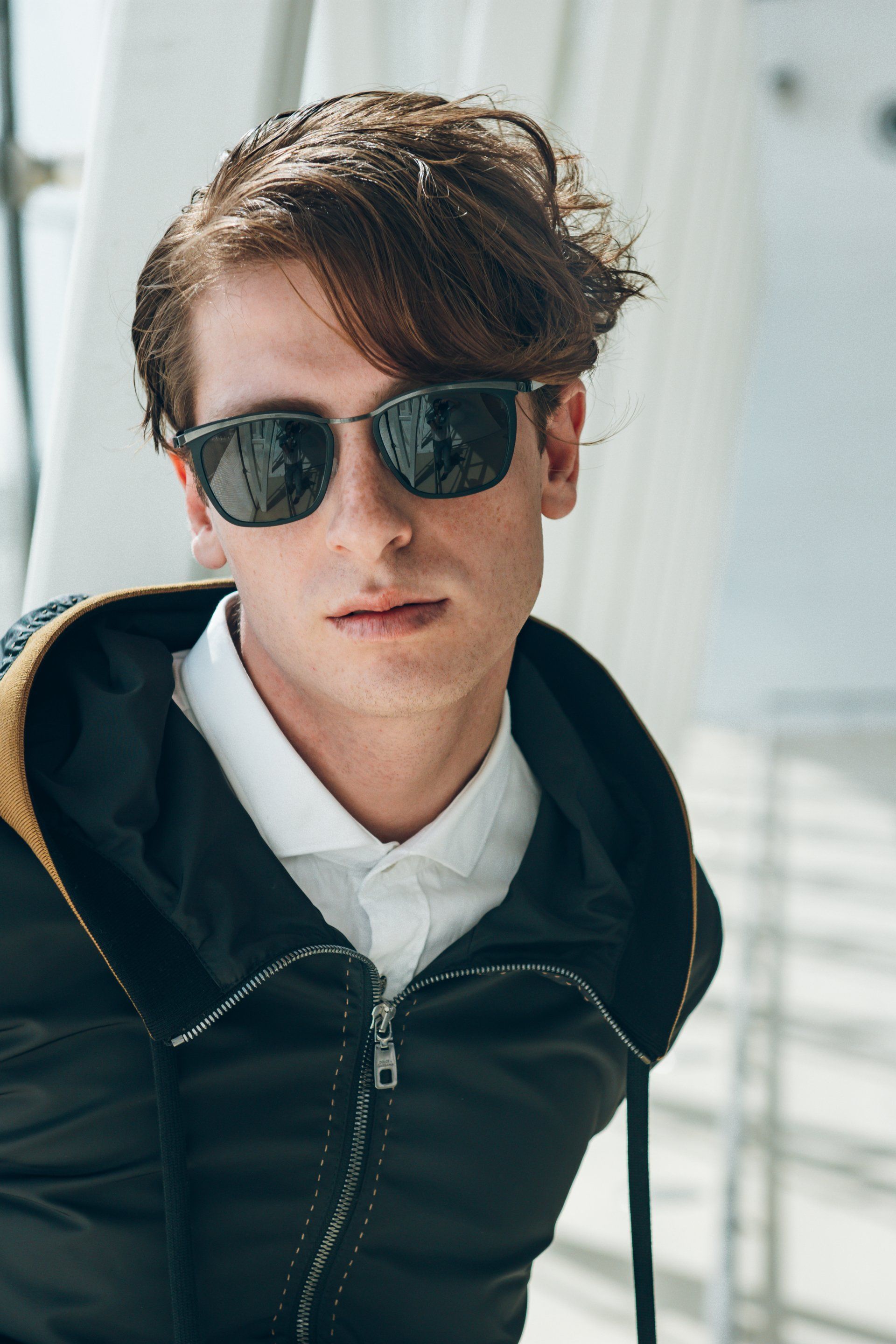 Mann mit Sonnenbrille, weißem Hemd und schwarzer Jacke, der vor einem weißen Hintergrund in die Kamera blickt.