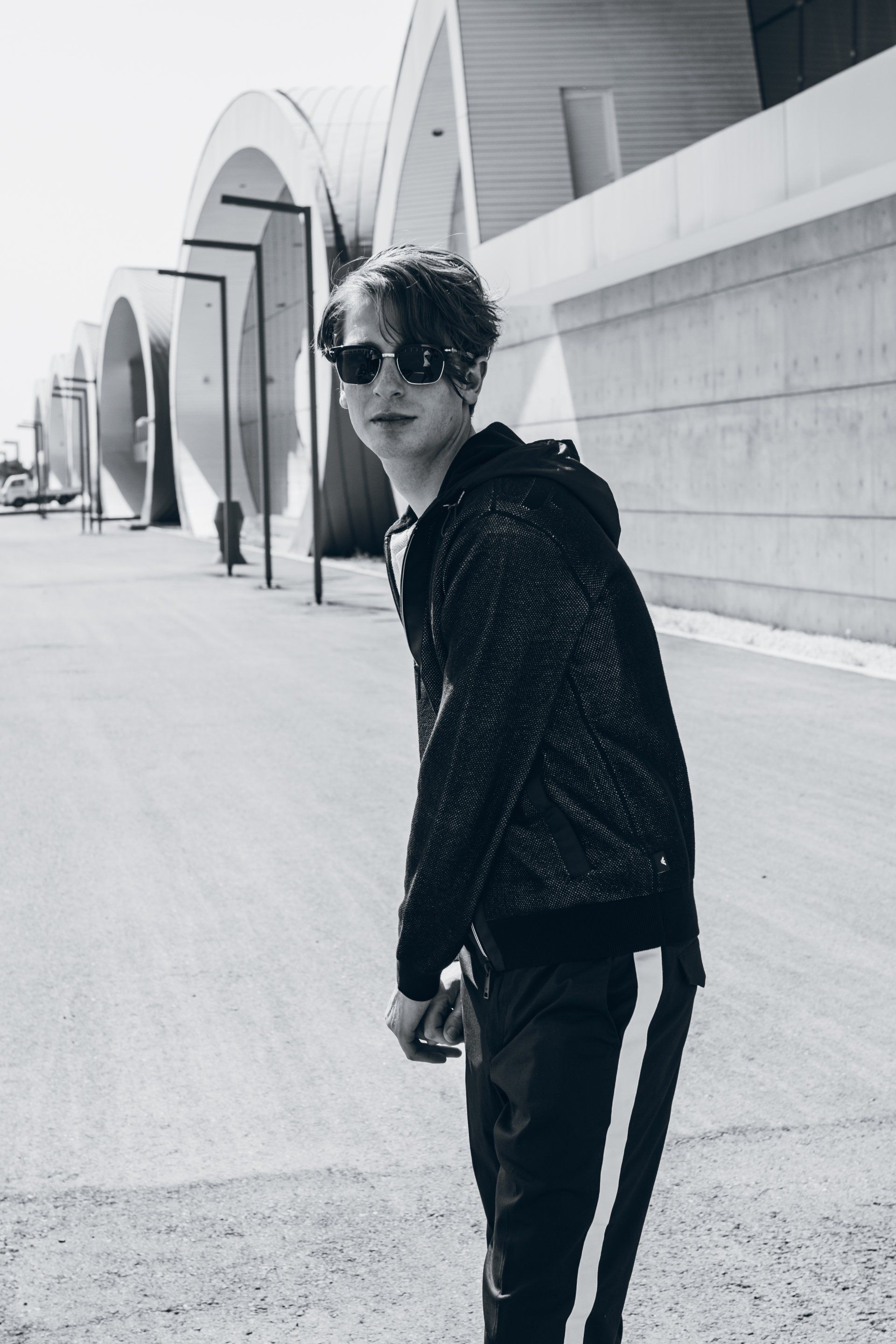 Ein junger Mensch mit Sonnenbrille und Kapuzenpullover steht auf einem Bürgersteig vor einem modernen Gebäude.