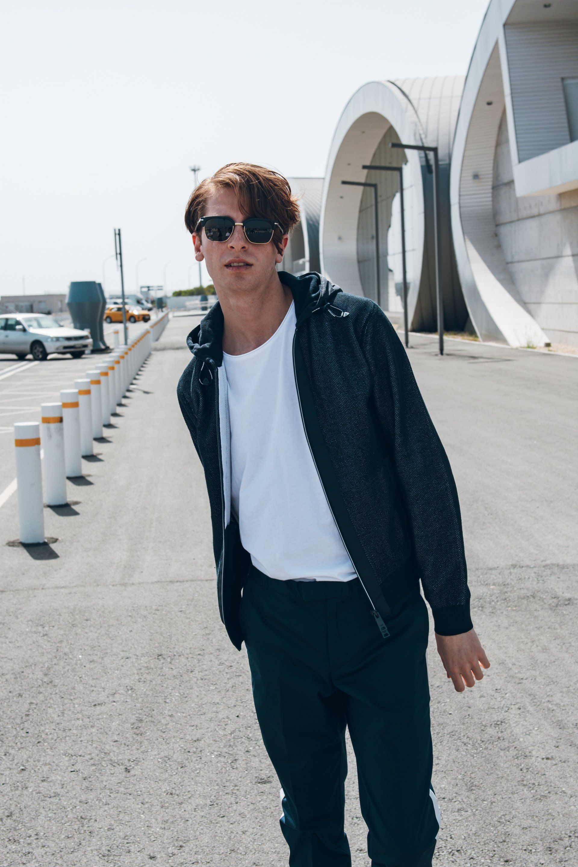 Mann mit Sonnenbrille und Jacke steht auf einer gepflasterten Fläche mit einem modernen Gebäude im Hintergrund.