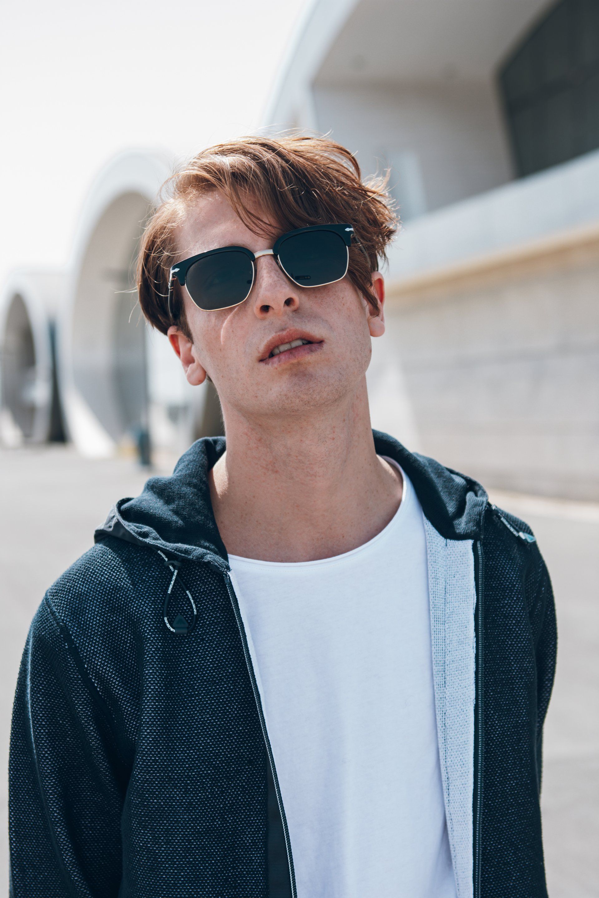 Mann mit Sonnenbrille und Kapuzenjacke steht im Freien in der Nähe moderner Architektur.