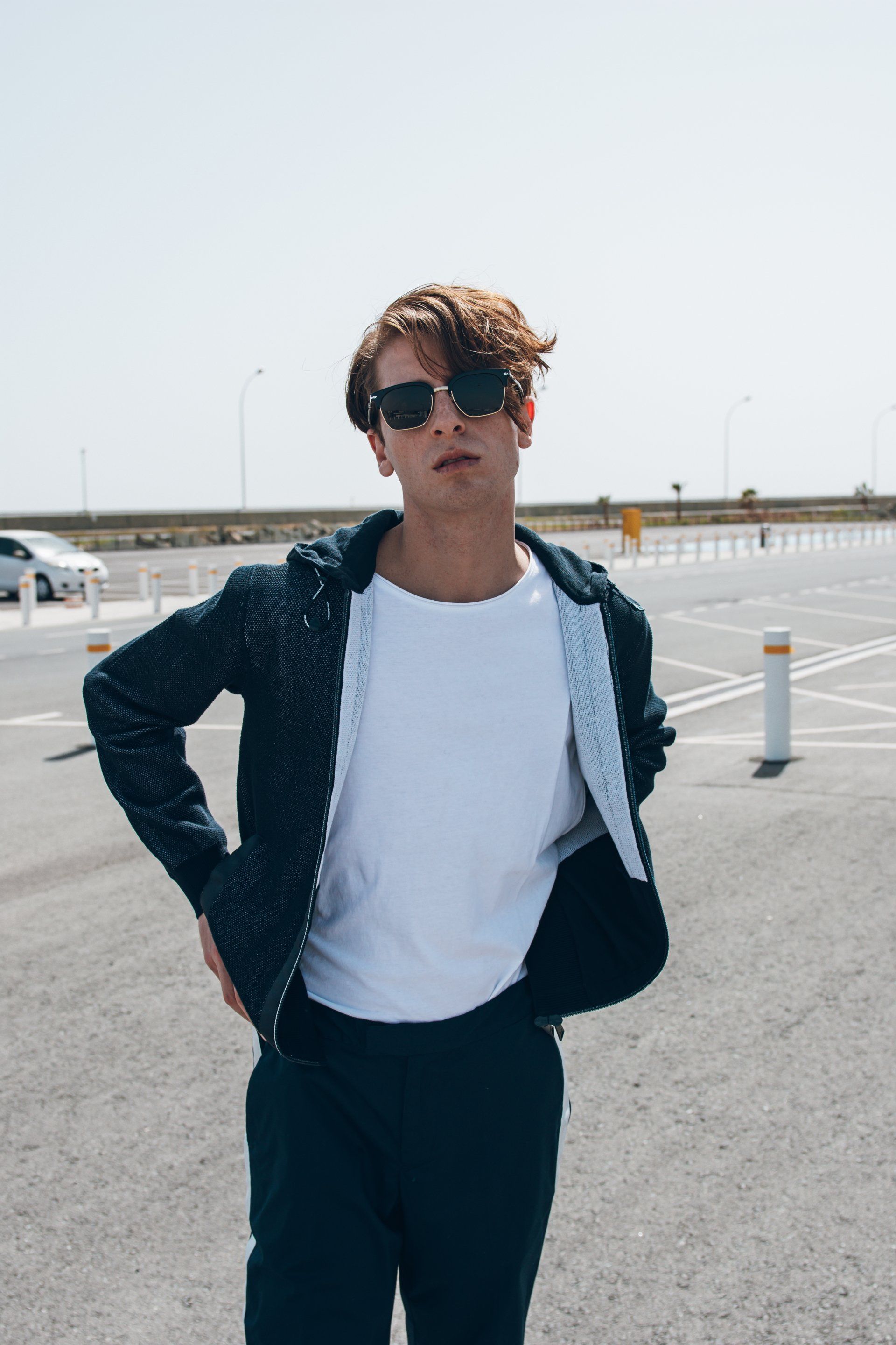 Mann mit Sonnenbrille auf einem Parkplatz, bekleidet mit Jacke und weißem Hemd, die Hände in die Hüften gestemmt.