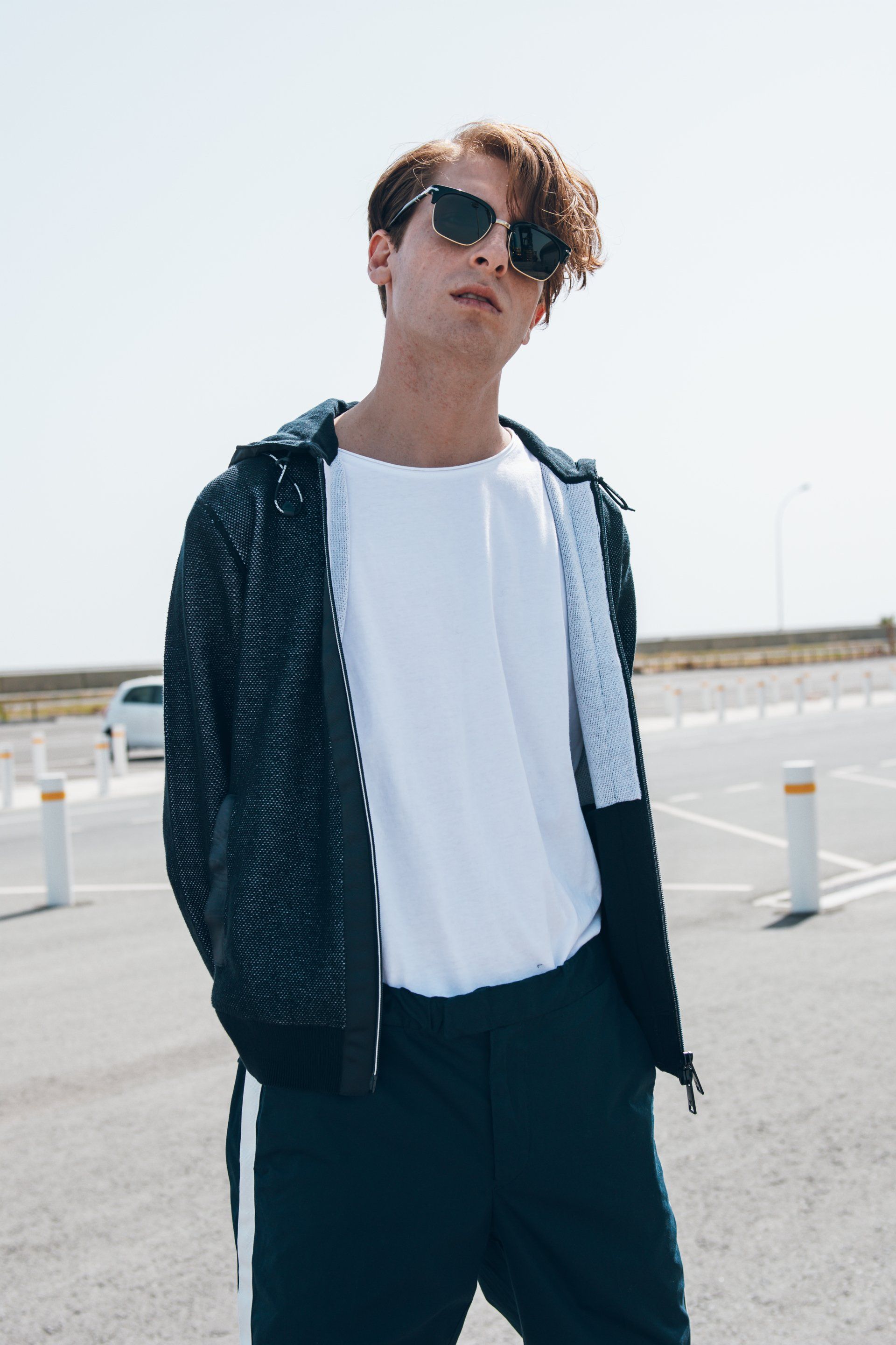 Mann mit Sonnenbrille, weißem T-Shirt und Trainingsanzug steht auf einem Parkplatz, posiert und blickt in die Kamera.