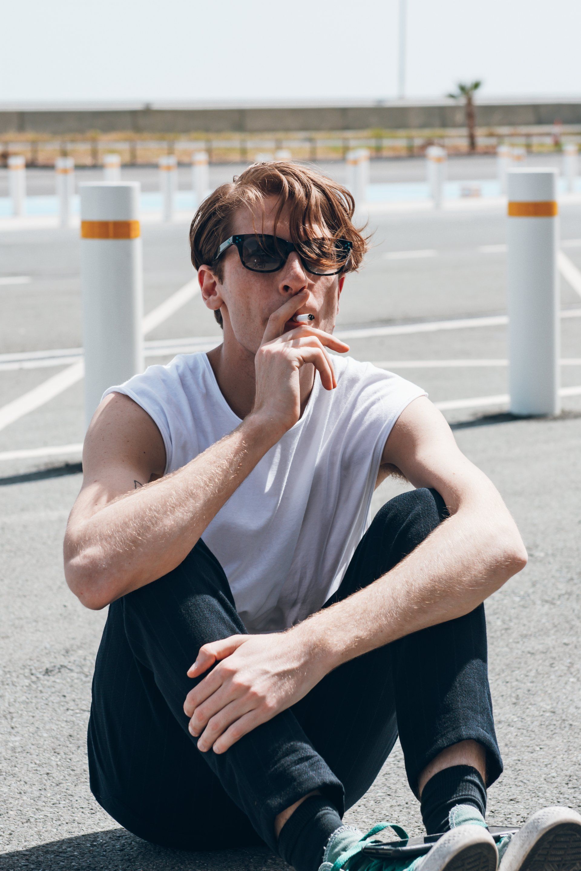 Mann mit Sonnenbrille sitzt auf Asphalt, Hand vor Gesicht, in der Nähe von weißen Pfosten.