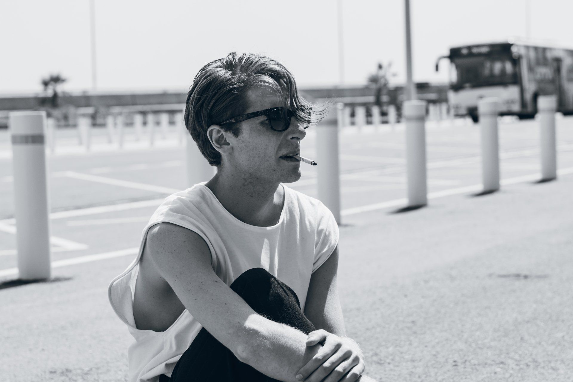 Mann mit Sonnenbrille raucht und sitzt an einem sonnigen Tag auf einem Parkplatz.