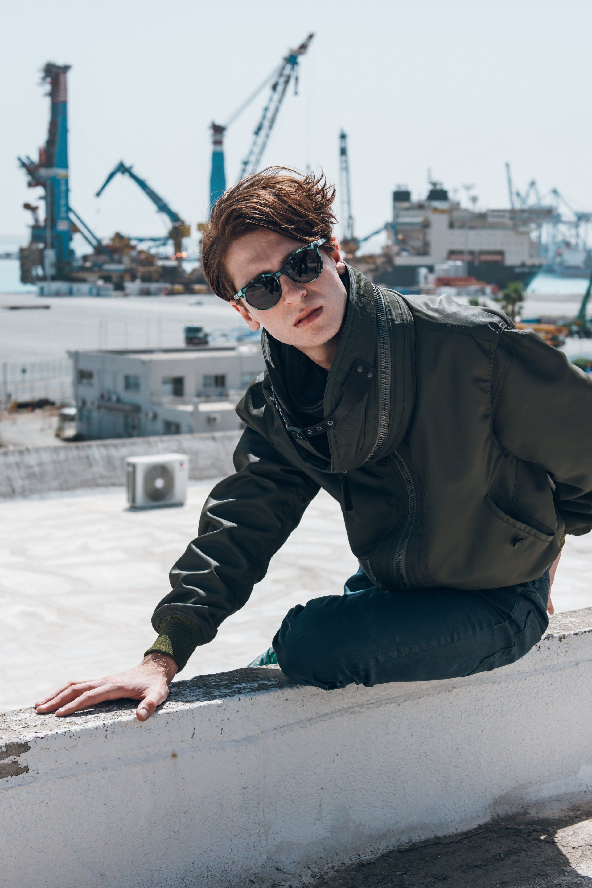 Mann mit Sonnenbrille und olivgrüner Jacke lehnt auf einem Dach, im Hintergrund der Hafen.