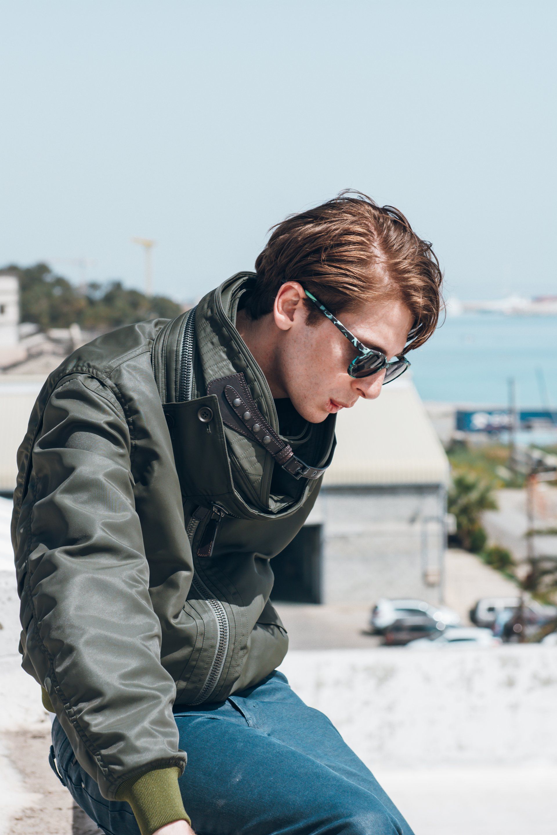 Mann in Fliegerjacke und Sonnenbrille, der im Freien an einer Wand lehnt.