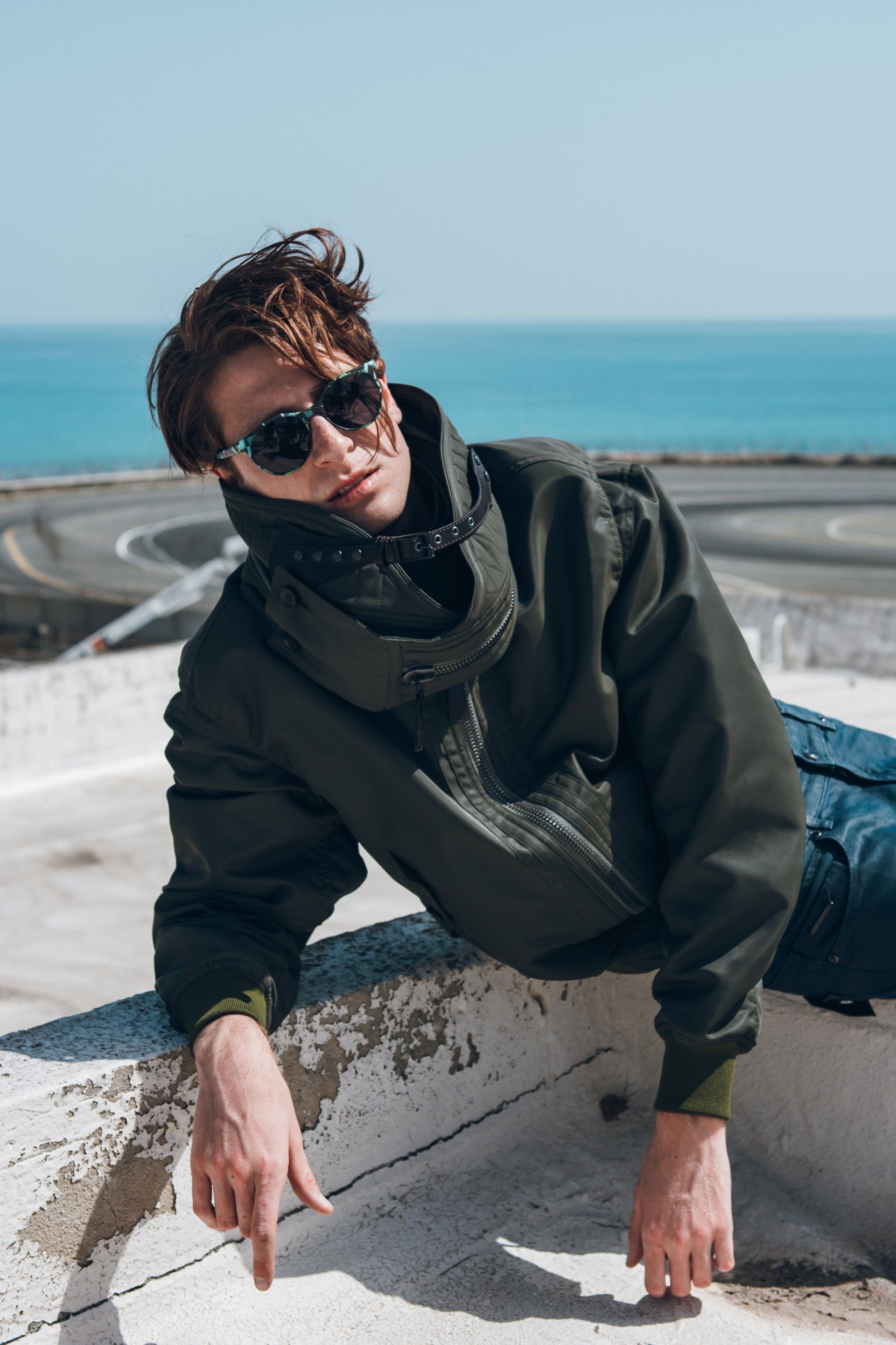 Mann mit Sonnenbrille, grüner Jacke und Jeans, liegt auf einem Dach mit Blick auf eine kurvenreiche Straße und das Meer.