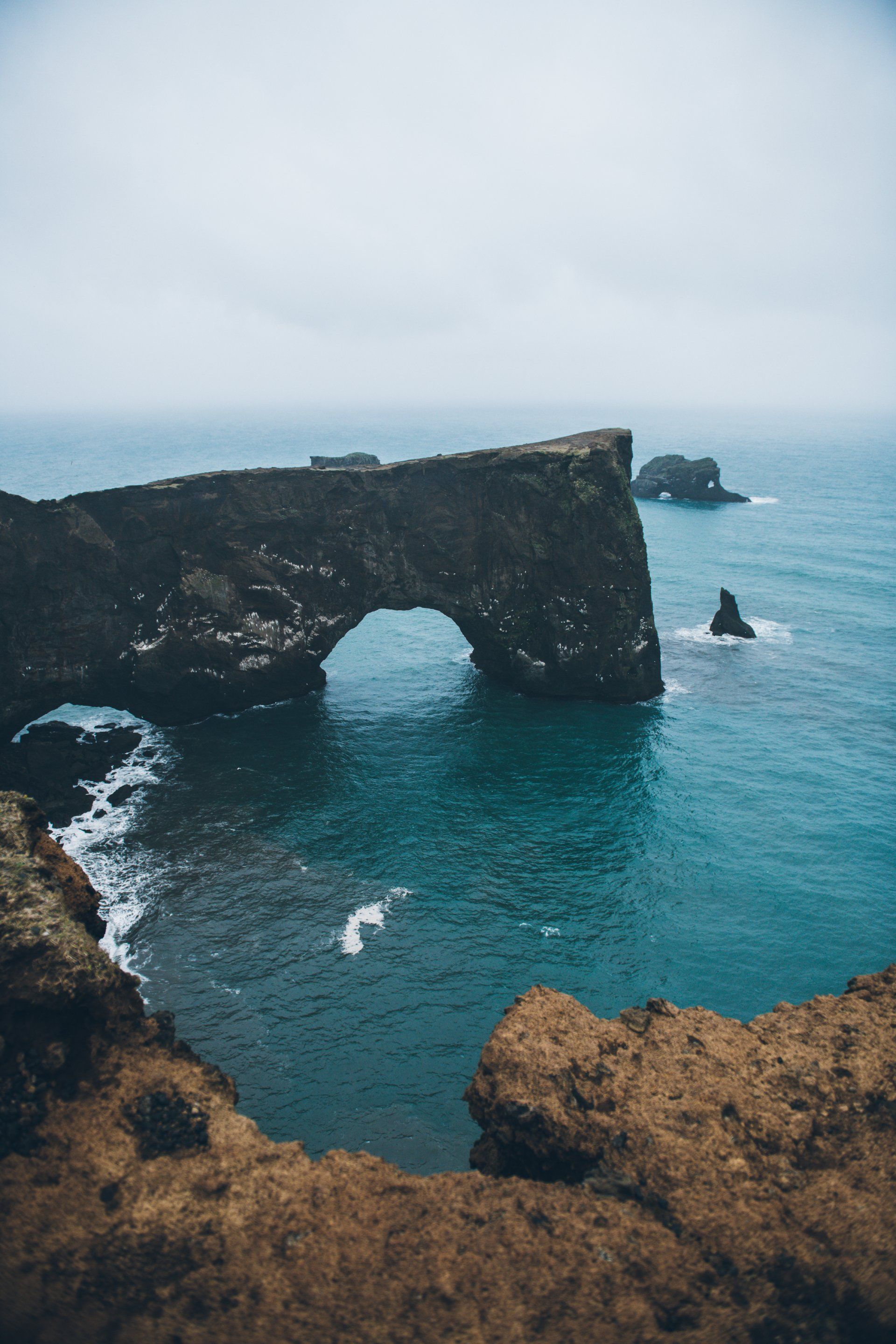 Eine bogenförmige Felsformation im blaugrünen Ozean unter einem bewölkten Himmel. Braune Klippen im Vordergrund.