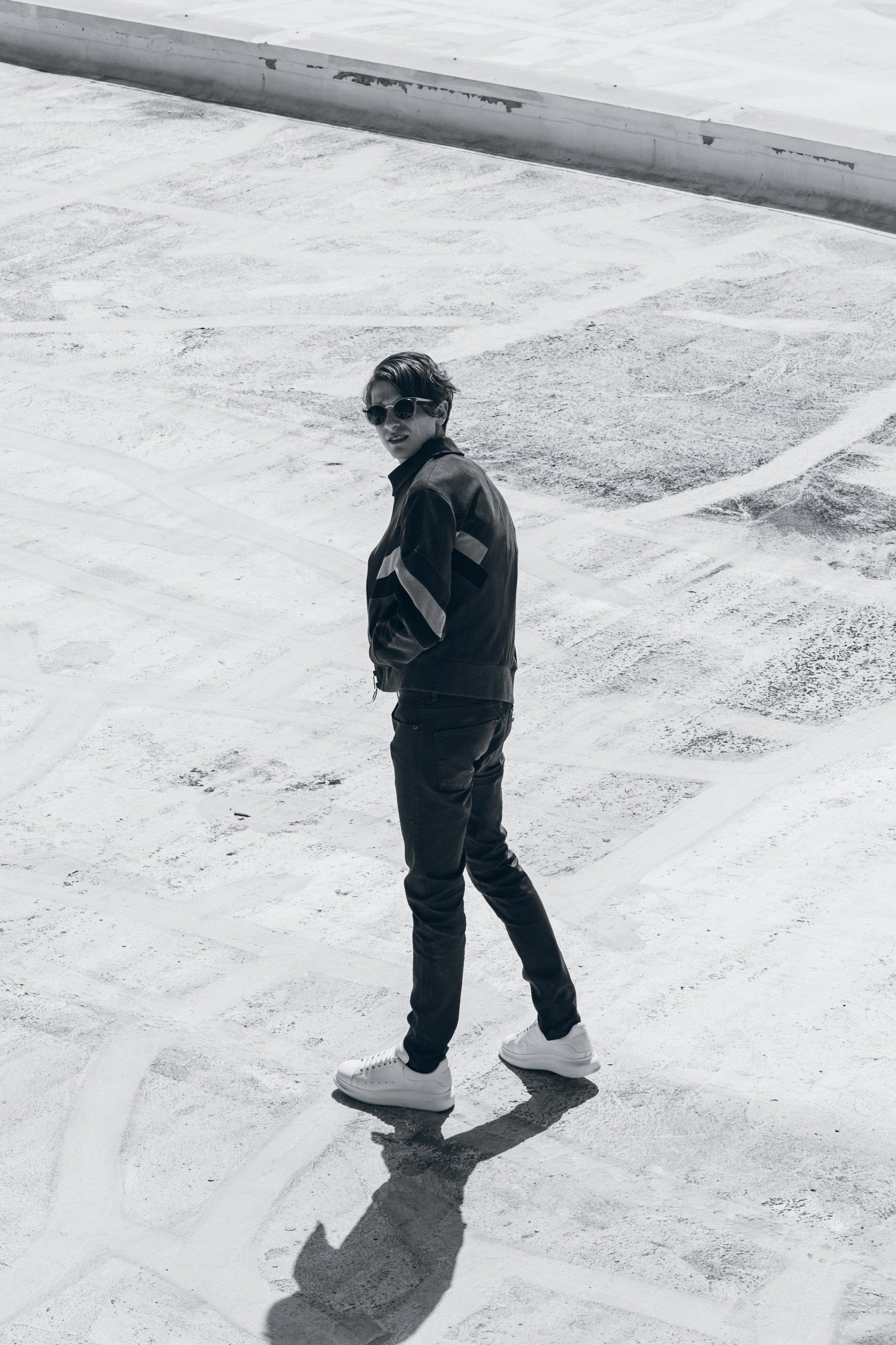 Mann in Jacke und Sonnenbrille schaut über die Schulter, steht auf hellem Untergrund.