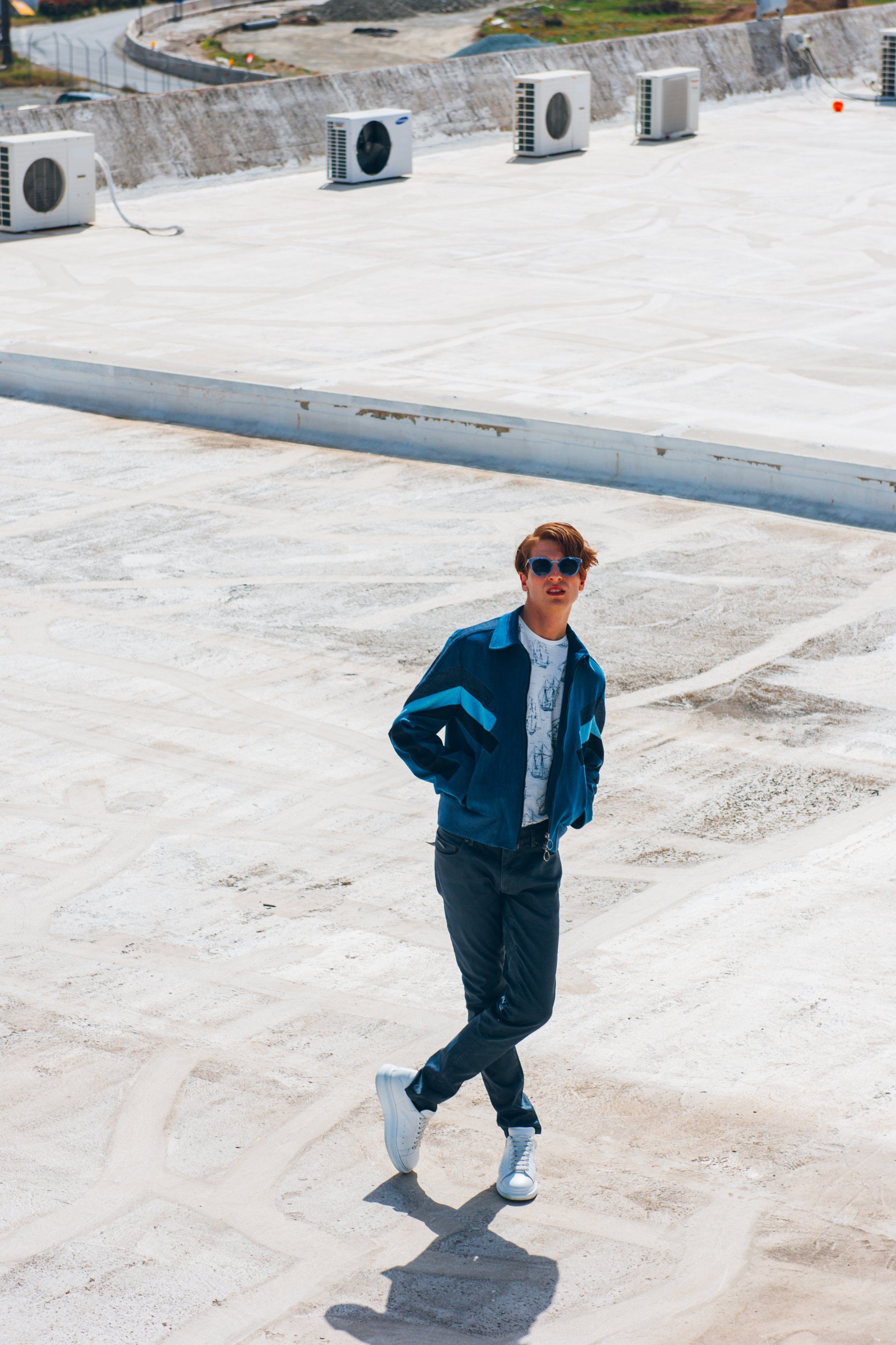 Mann in blauer Jacke, Sonnenbrille und weißen Turnschuhen posiert auf einem sonnigen Dach.