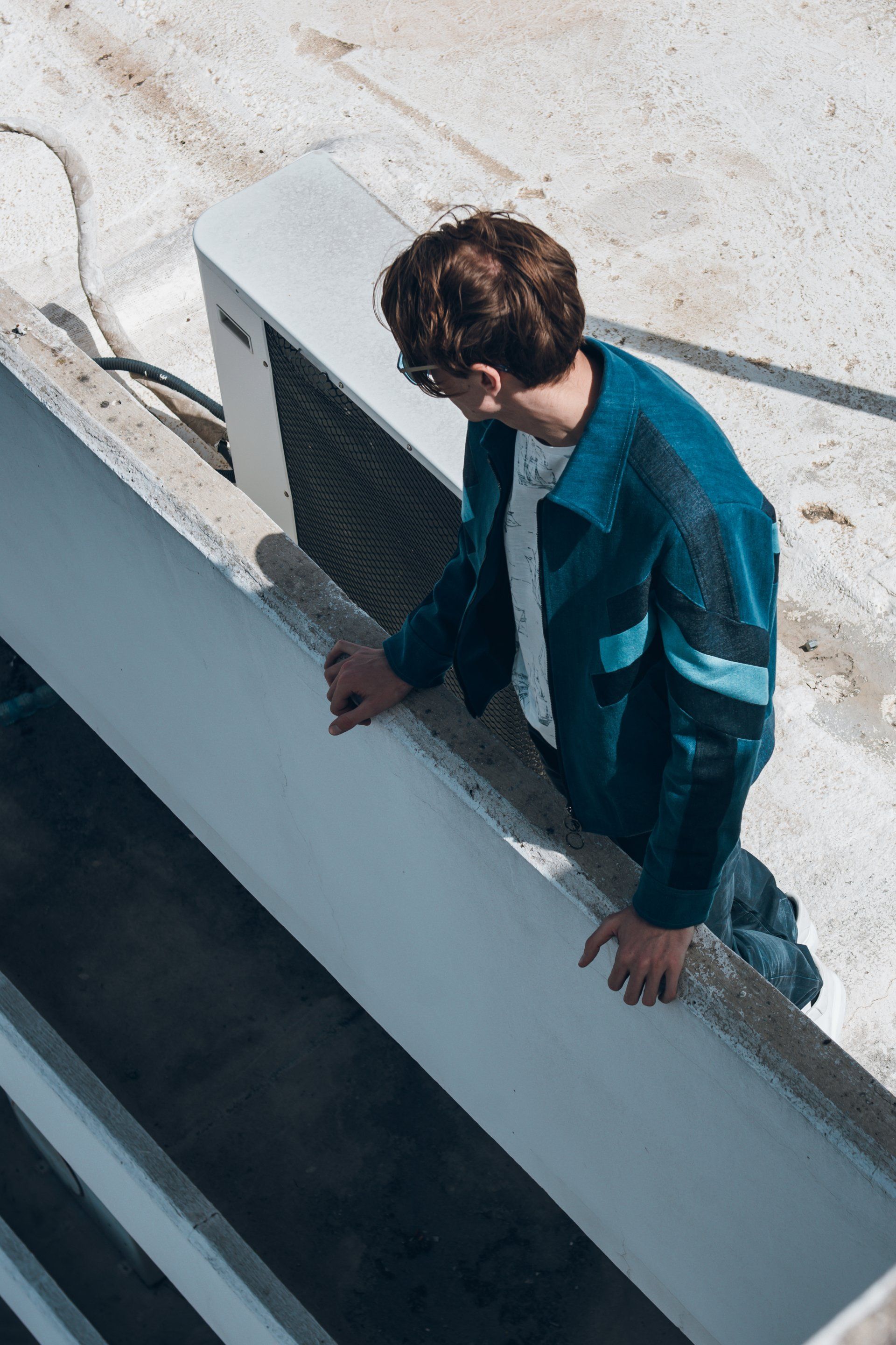 Mann in blauer Jacke lehnt auf einem Betonvorsprung und blickt nach unten. Sonnenlicht auf dem Dach.