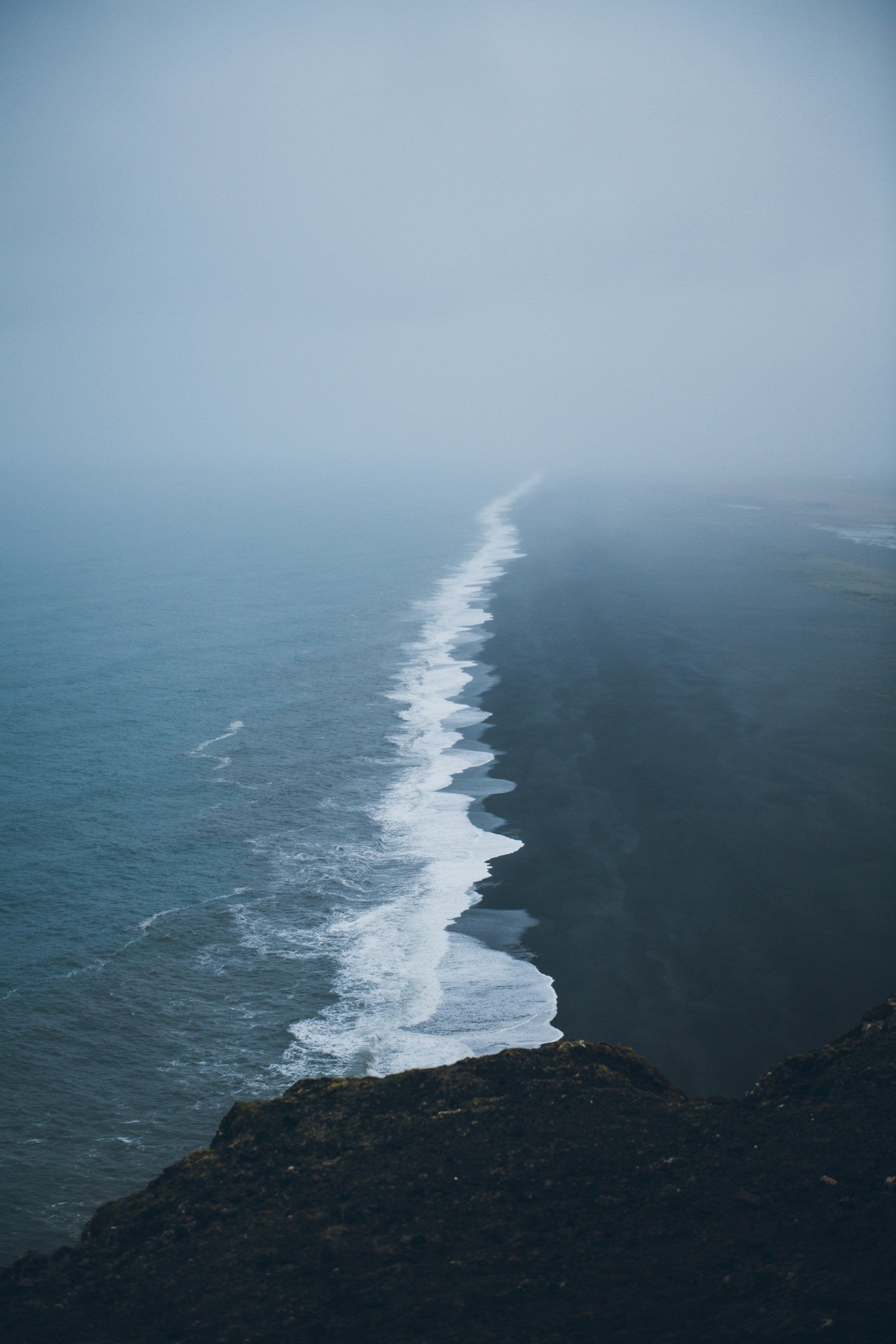 Schwarzer Sandstrand trifft auf stürmischen Ozean, in Nebel gehüllt unter bewölktem Himmel.