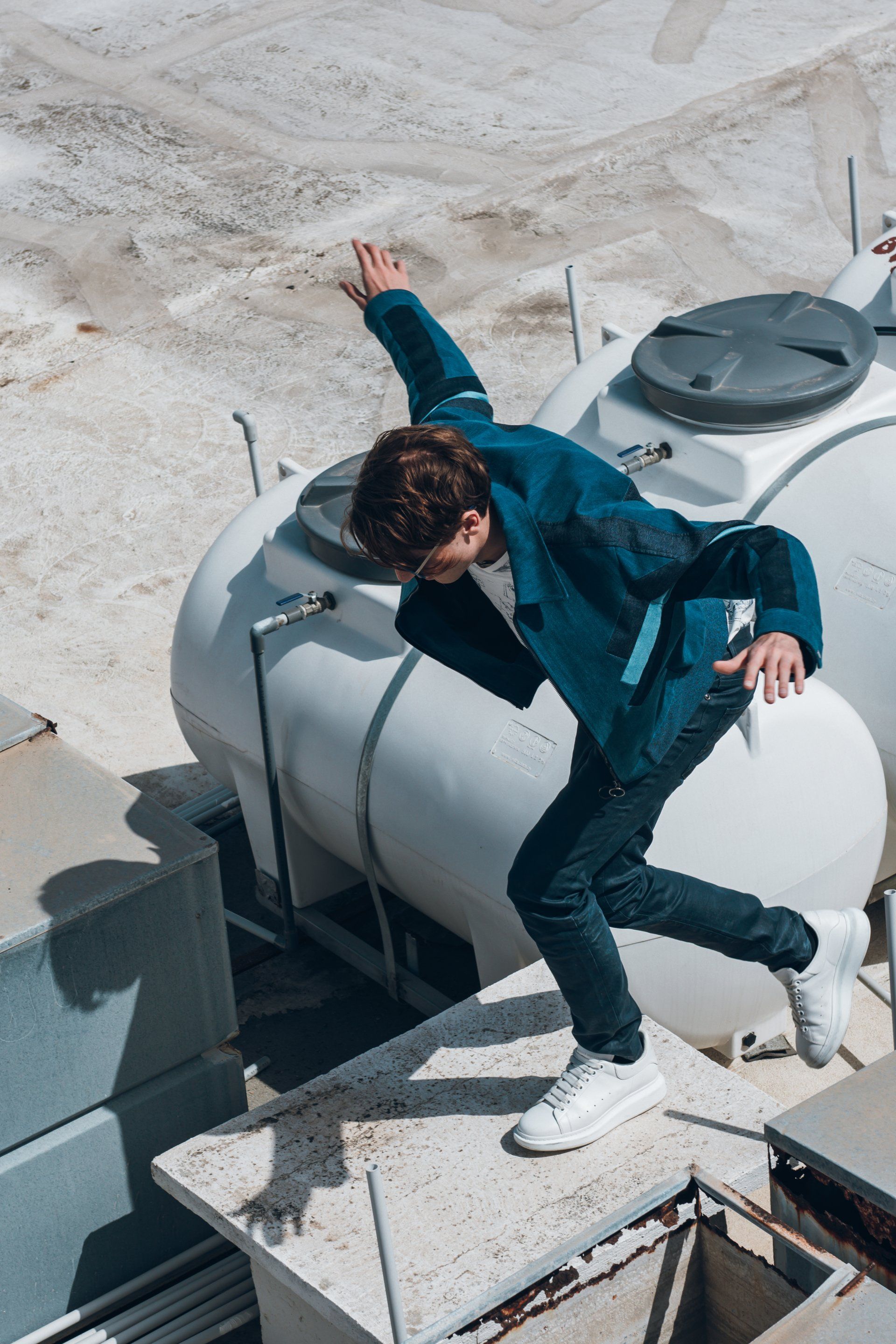 Mann im blauen Blazer springt von einem Dach, weiße Panzer im Hintergrund, sonniger Tag.