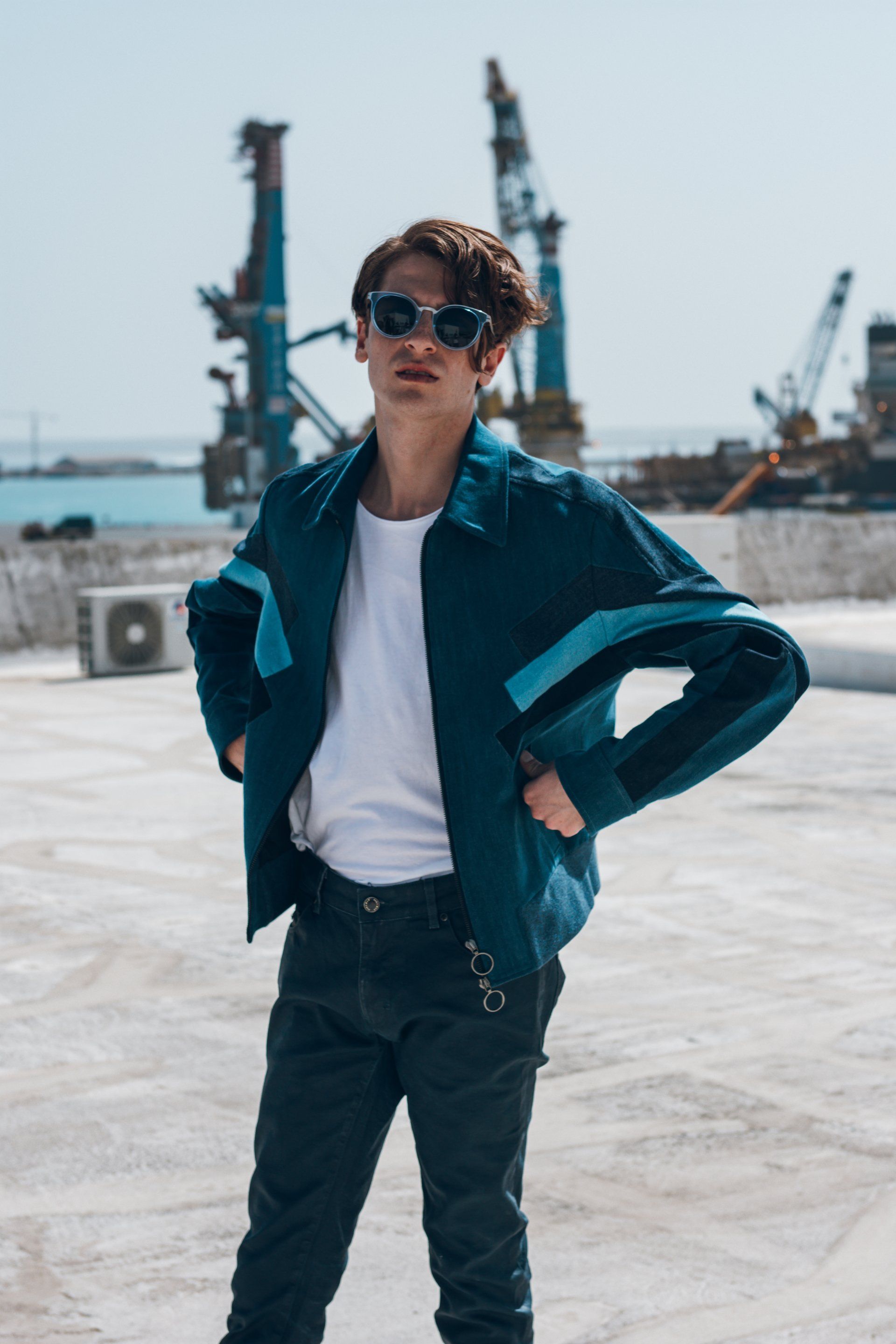 Person in blauer Jacke und Sonnenbrille steht mit den Händen in den Hüften auf einem Dach, im Hintergrund Industriekräne.