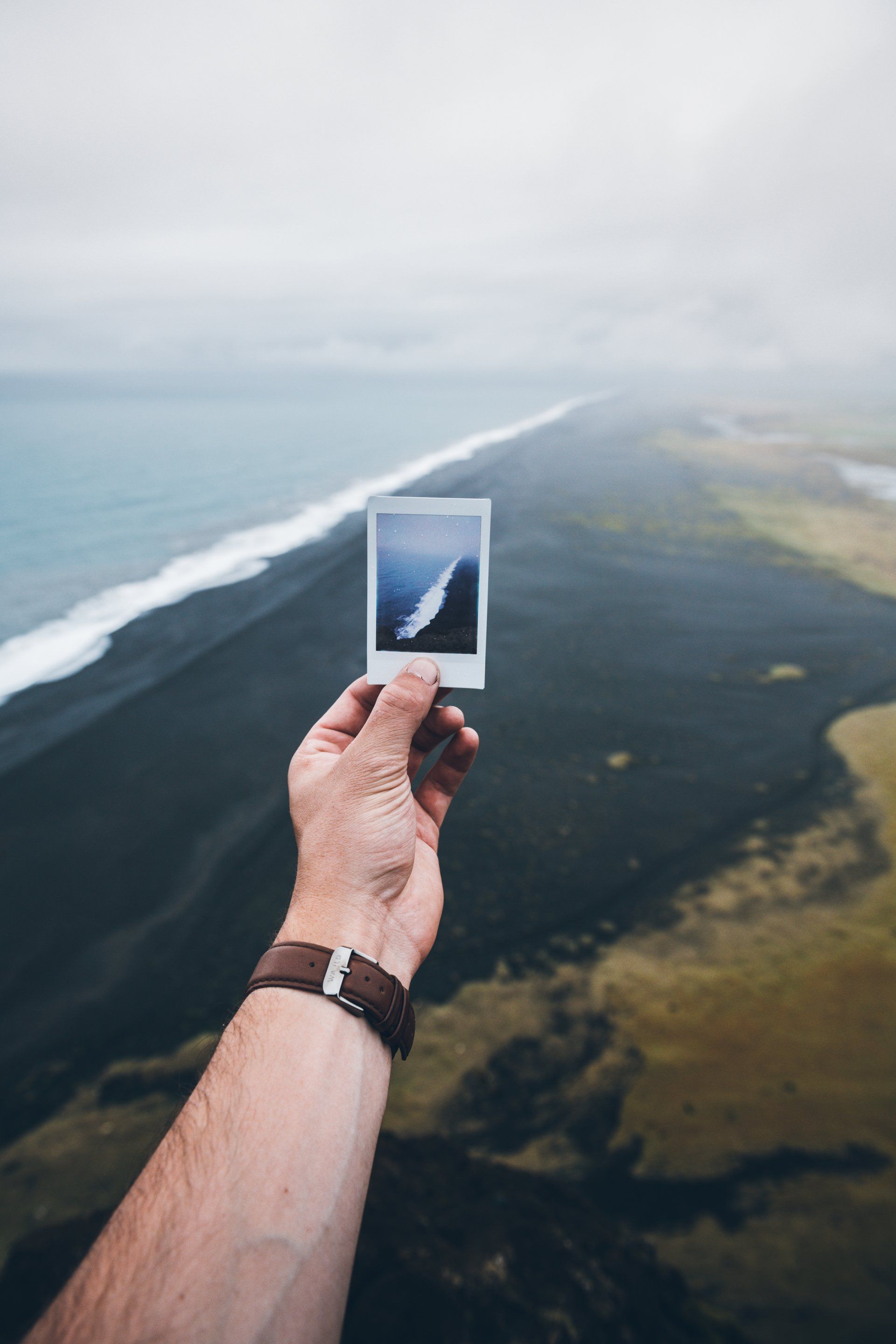 Hand hält Polaroid einer Küstenlandschaft. Schwarzer Sandstrand, blaues Meer und bewölkter Himmel.