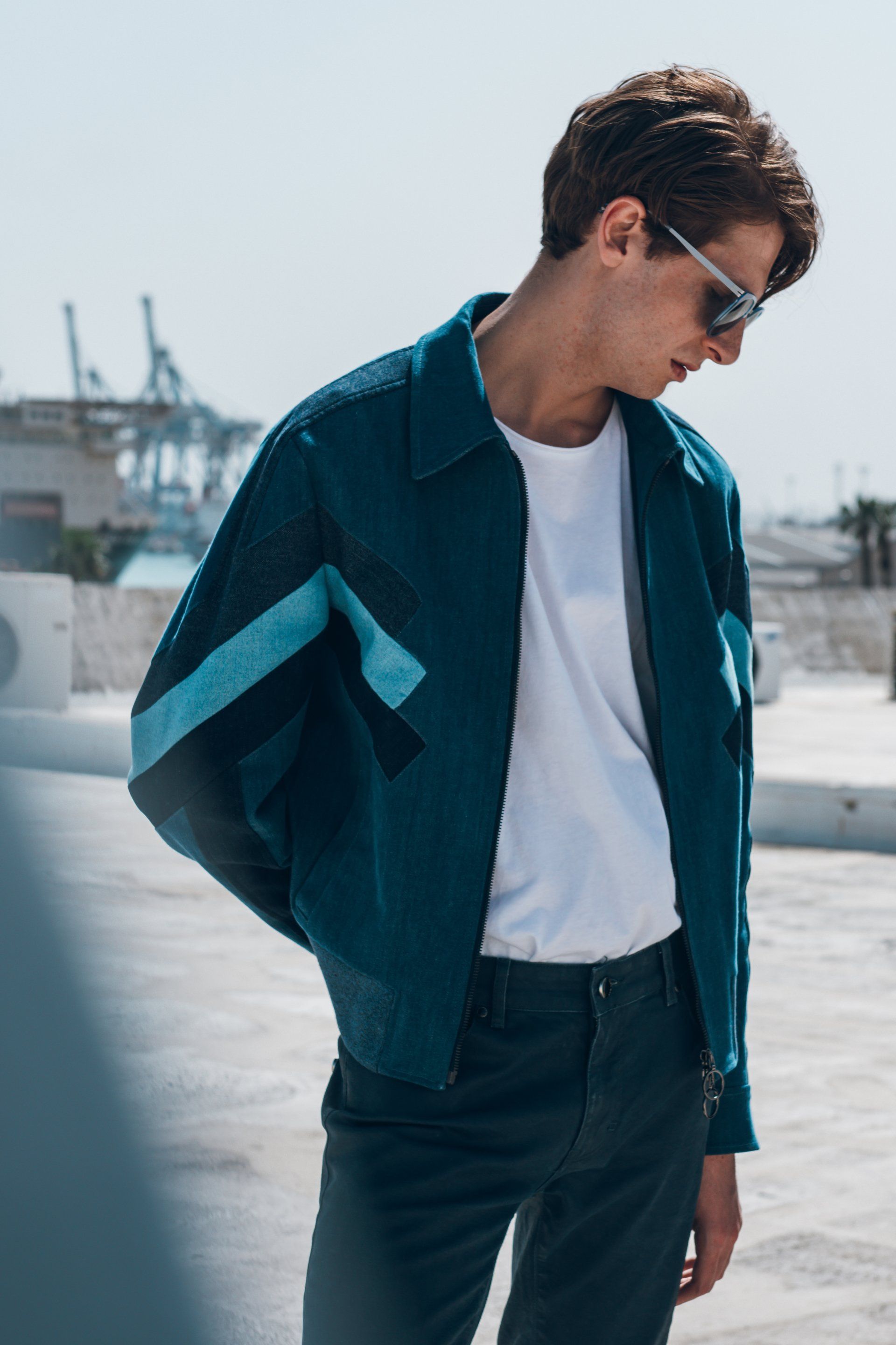 Mann mit Sonnenbrille, blauer Jacke und weißem T-Shirt steht auf einem Dach.