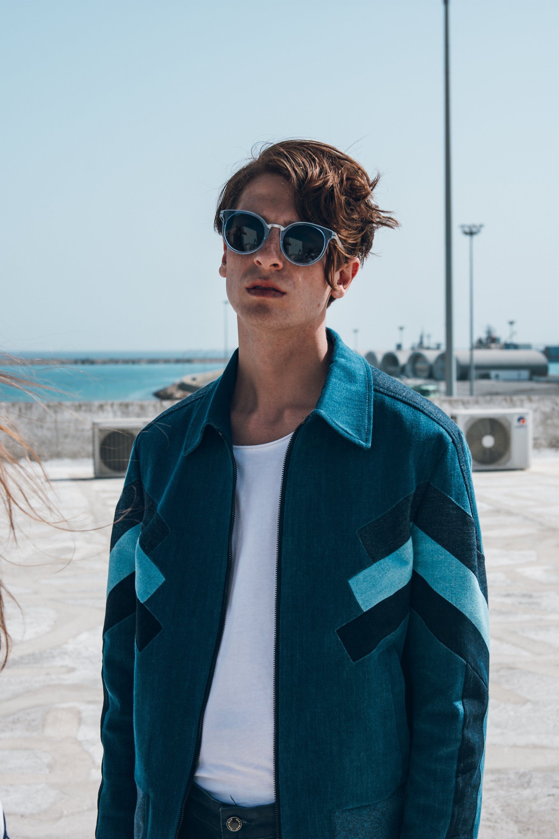 Junger Mann mit Sonnenbrille und blauer Jacke steht draußen am Meer.