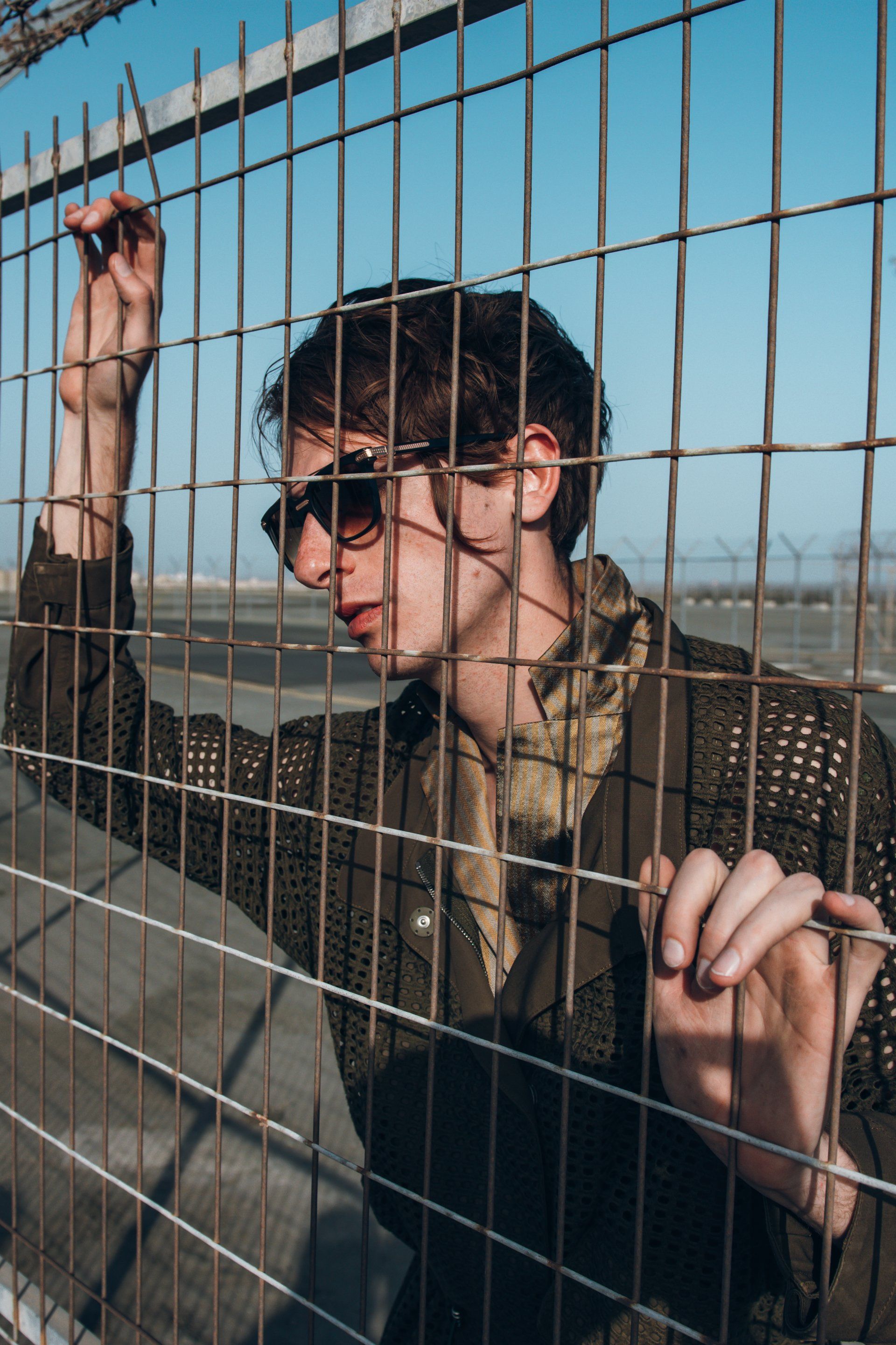 Mann mit Sonnenbrille lehnt im Freien an einem Metallzaun und trägt eine gemusterte Jacke unter blauem Himmel.