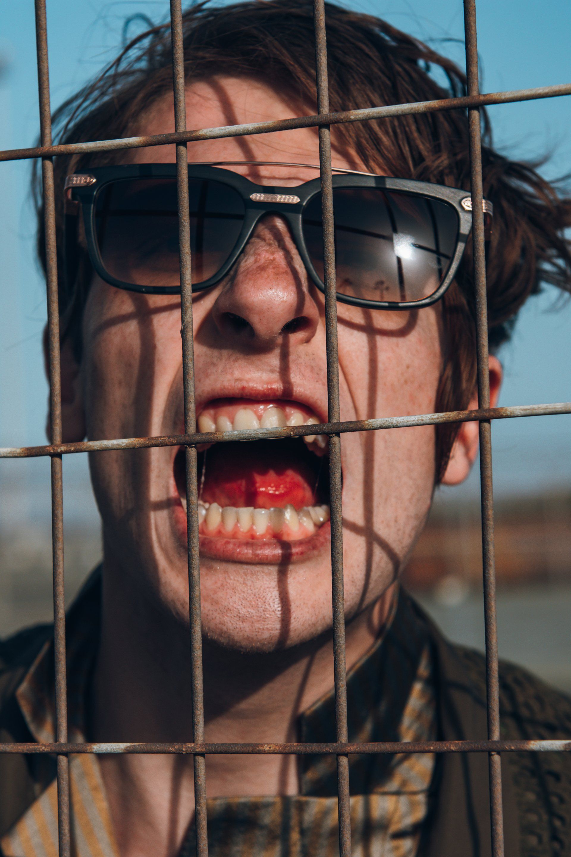 Mann mit Sonnenbrille schreit mit offenem Mund hinter einem rostigen Zaun.