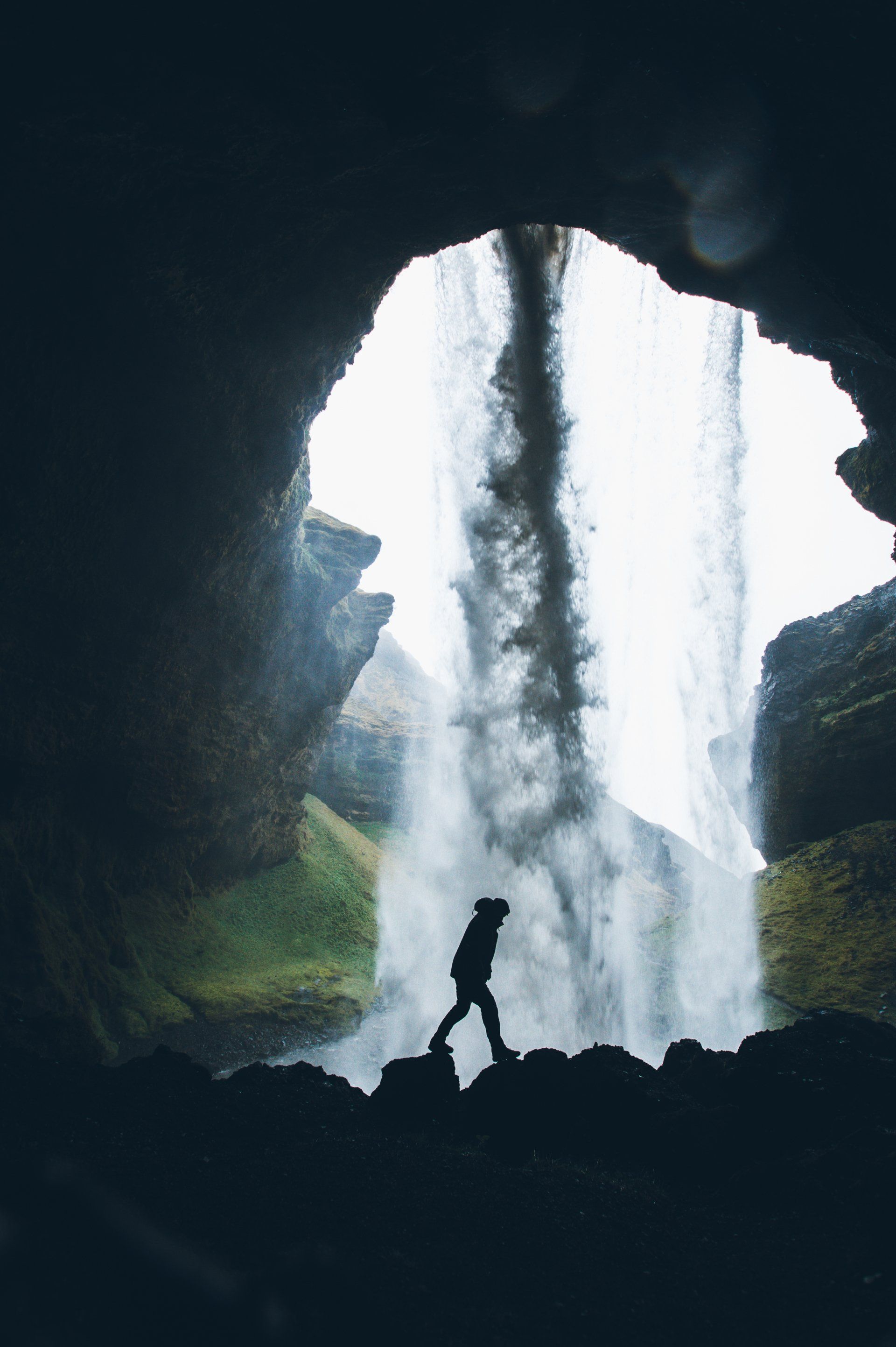 Silhouette einer Person, die vor einem Wasserfall in einer Höhle geht; umgeben von hellem Licht und Nebel.