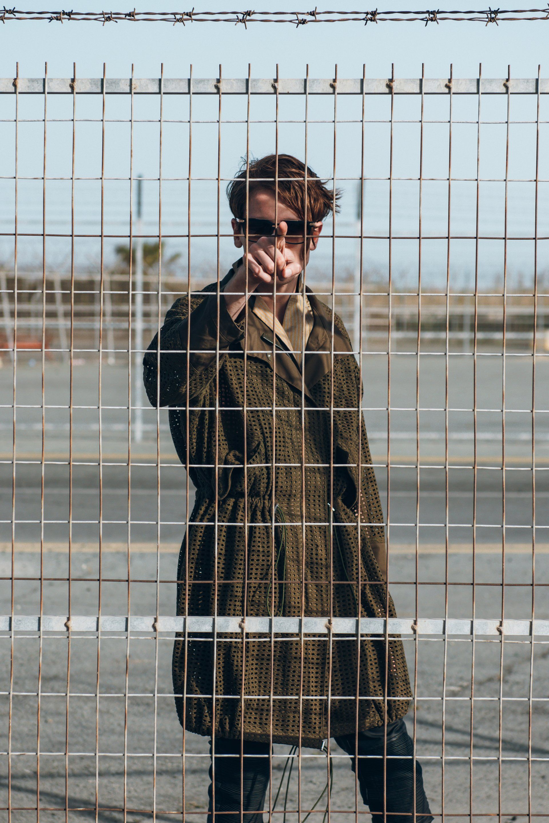 Person mit Sonnenbrille und gemustertem Mantel, die hinter einem Metallzaun steht.