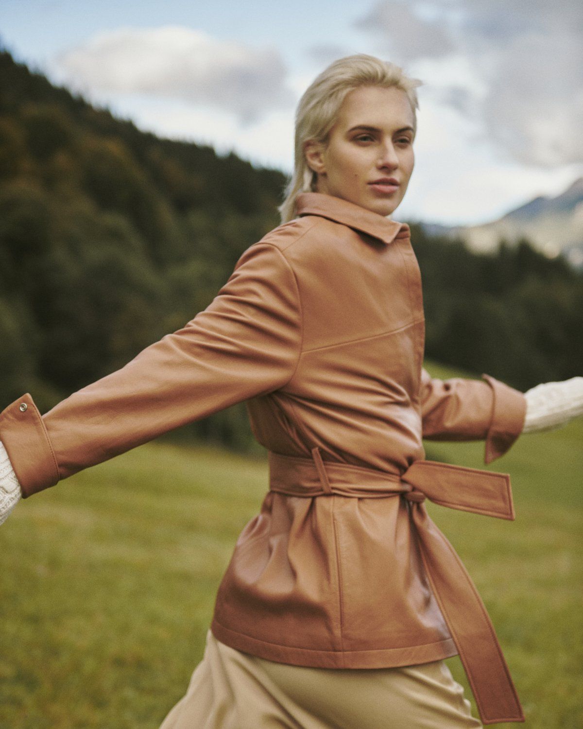 Frau in hellbrauner Jacke und Hose, die Arme auf einer grünen Wiese mit Bergen im Hintergrund ausgestreckt.