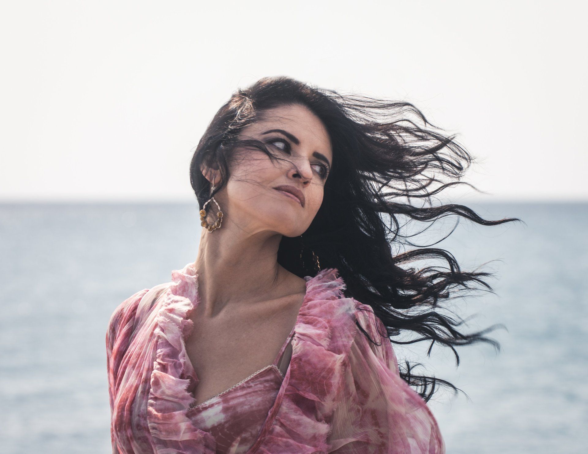 Frau mit dunklem Haar, das im Wind weht, blickt nach oben und trägt ein rosa Kleid am Meer.