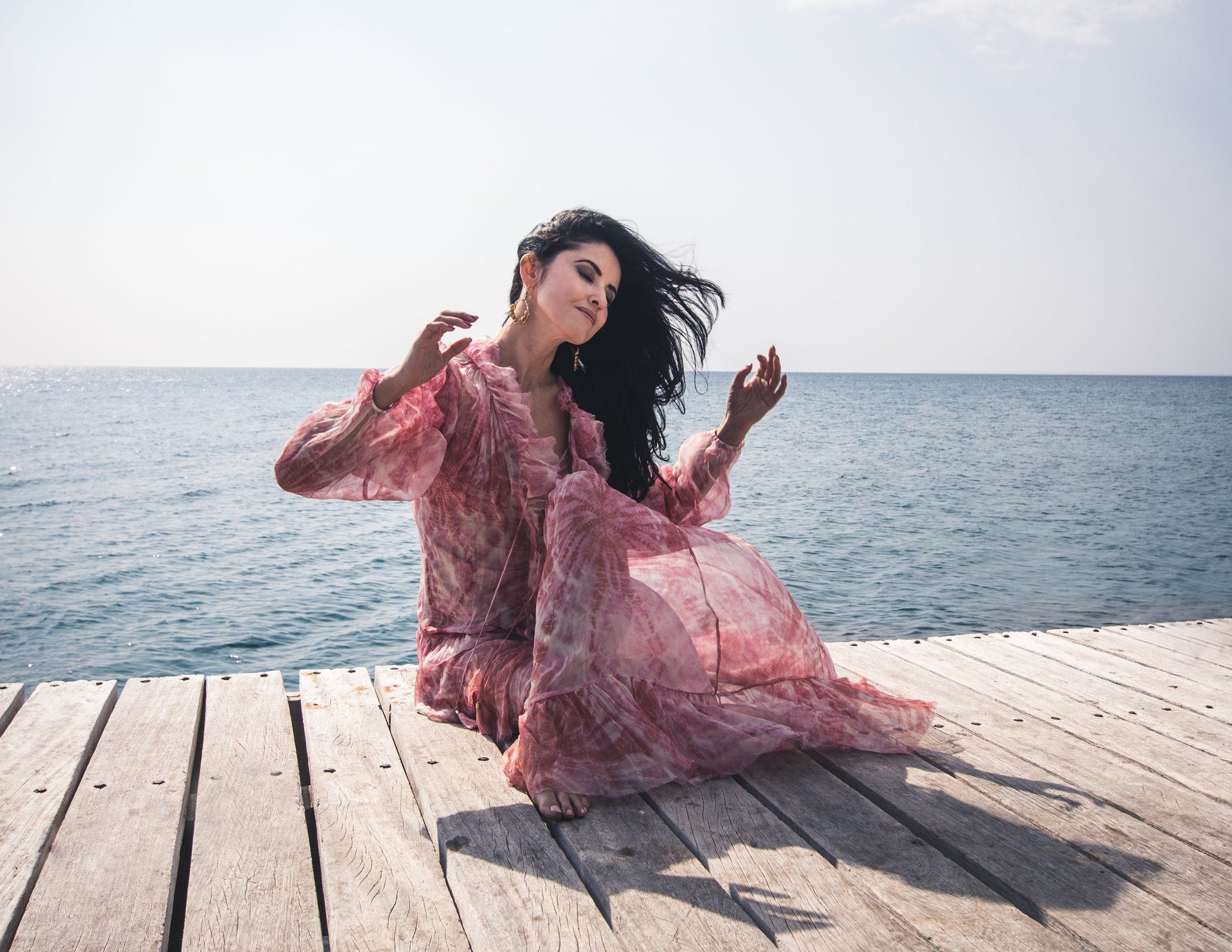 Eine Frau im rosa Kleid sitzt auf einem Holzsteg am Meer, ihr Haar weht im Wind.