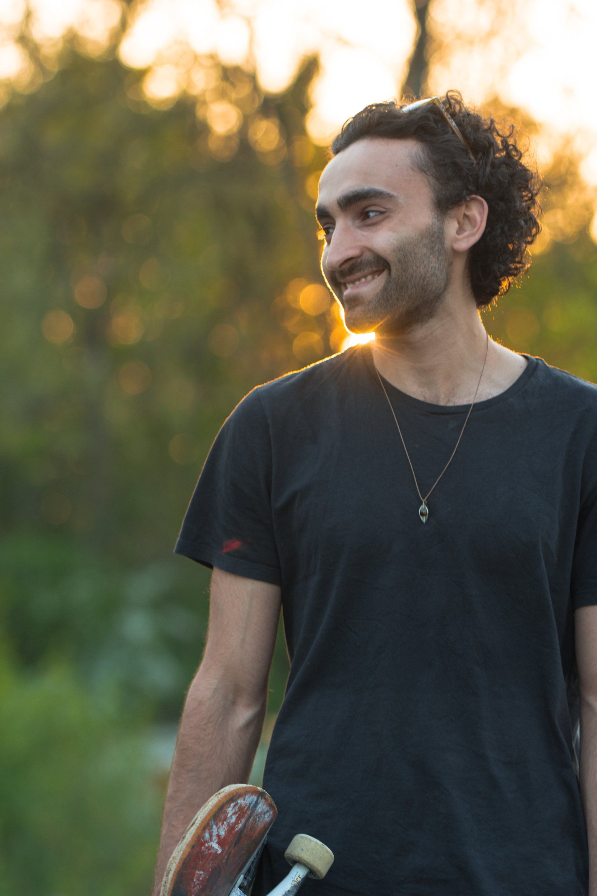 Mann mit lockigem Haar lächelt, während er bei Sonnenuntergang in einem Park ein Skateboard hält.