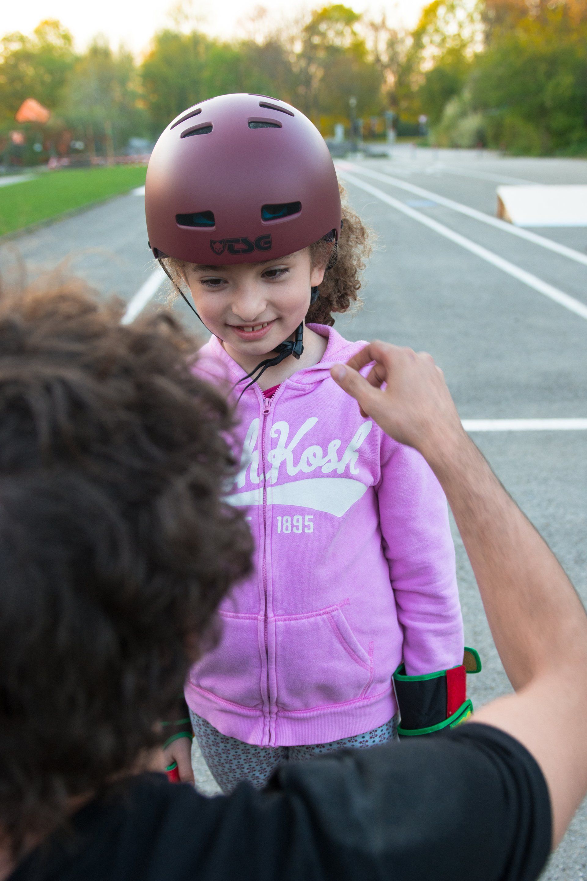 Junges Mädchen in rosa Kapuzenpullover und Helm lächelt, während Erwachsene ihren Helm zurechtrückt.