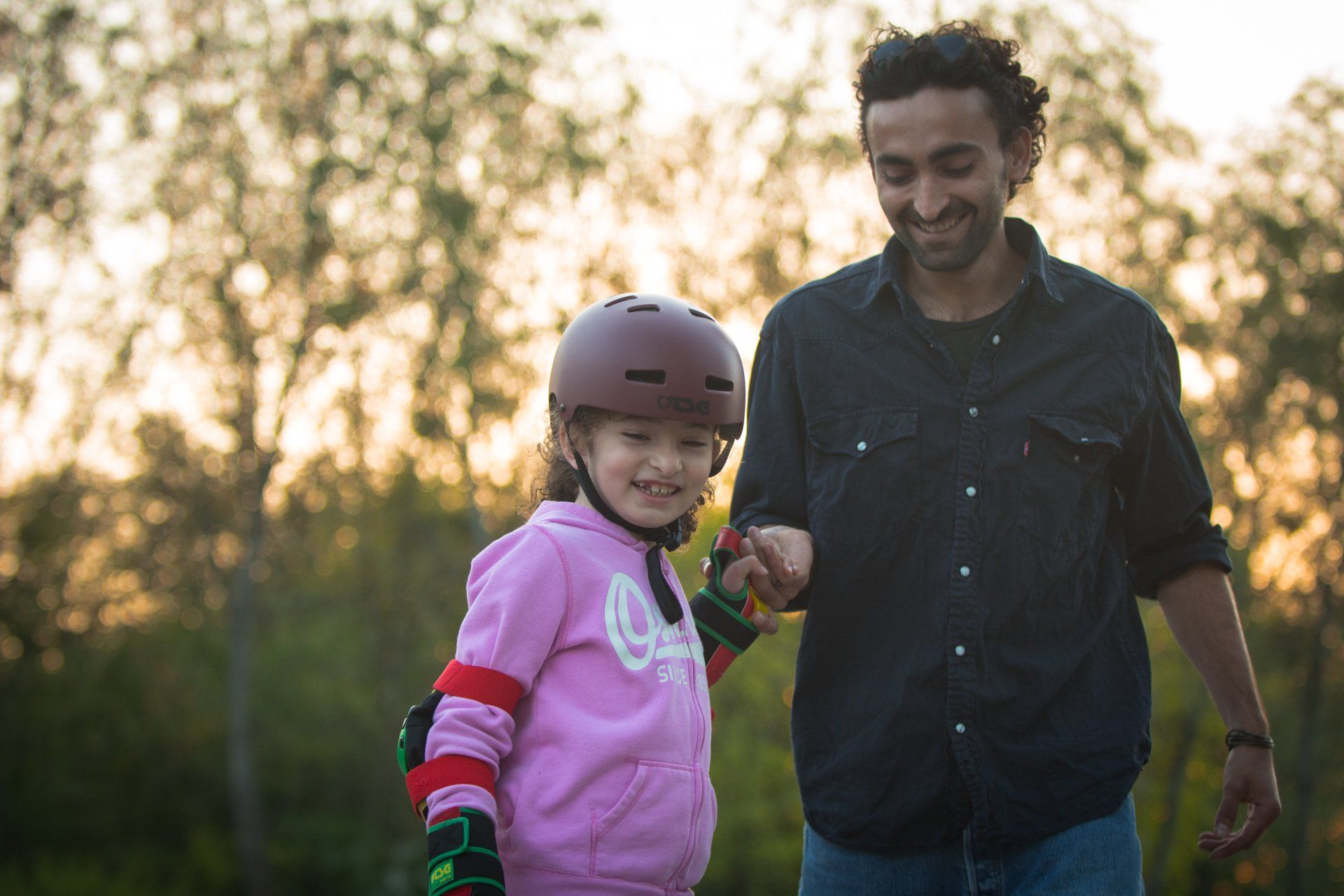 Vater hilft seinem Kind, Helm und Rollschuh anzuziehen, draußen. Lächelnd, goldenes Licht.